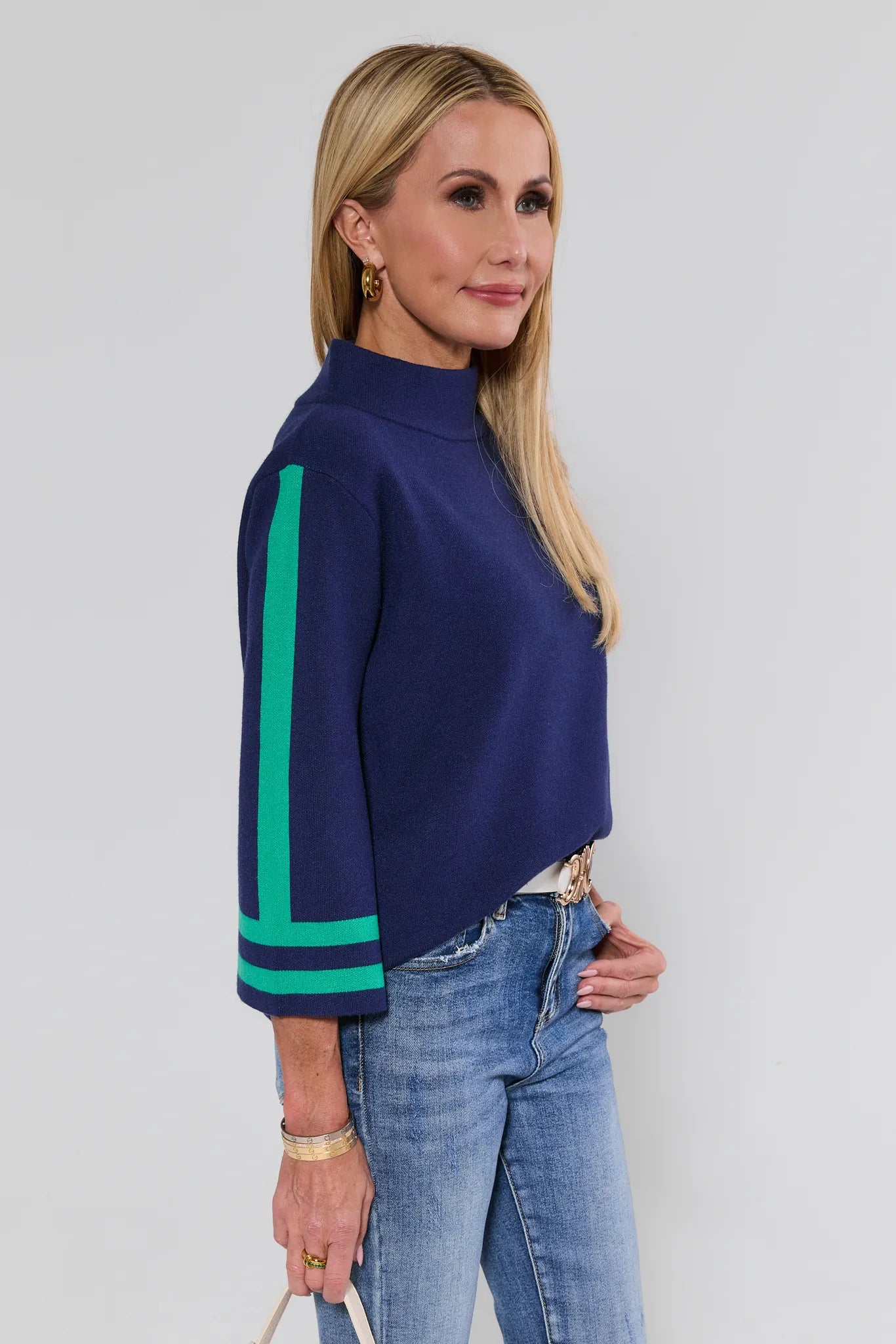 Woman wearing a navy sweater with green accents and blue jeans against a plain background