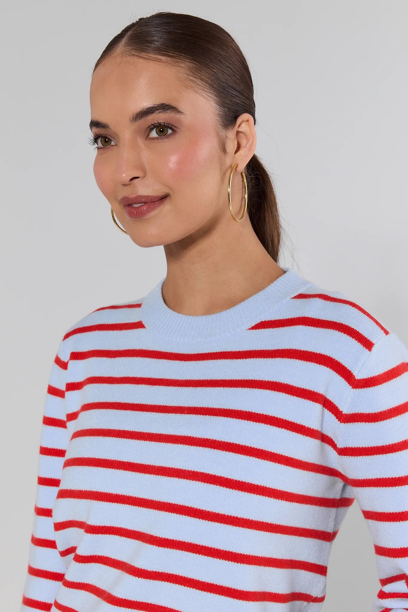 Woman wearing a red and white striped sweater and gold earrings with blue jeans on a plain background