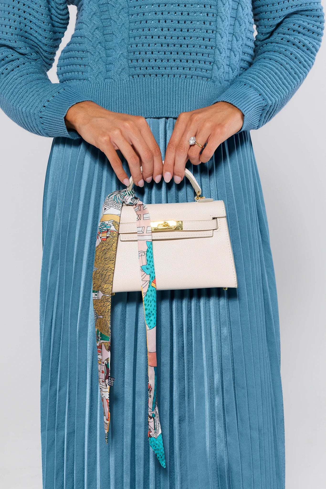 Person wearing a blue sweater and pleated skirt holding a white handbag with a colorful strap.