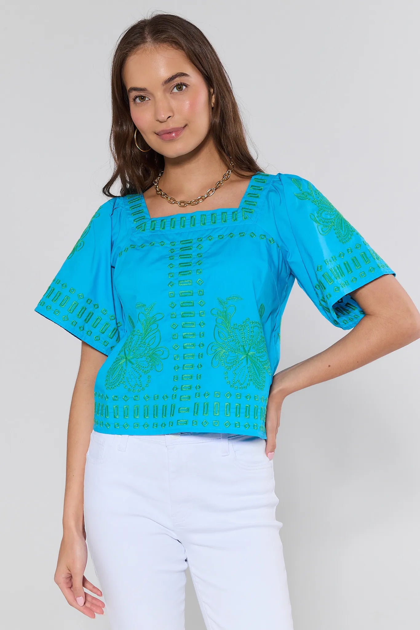 Woman wearing a blue embroidered top and white pants on a plain background