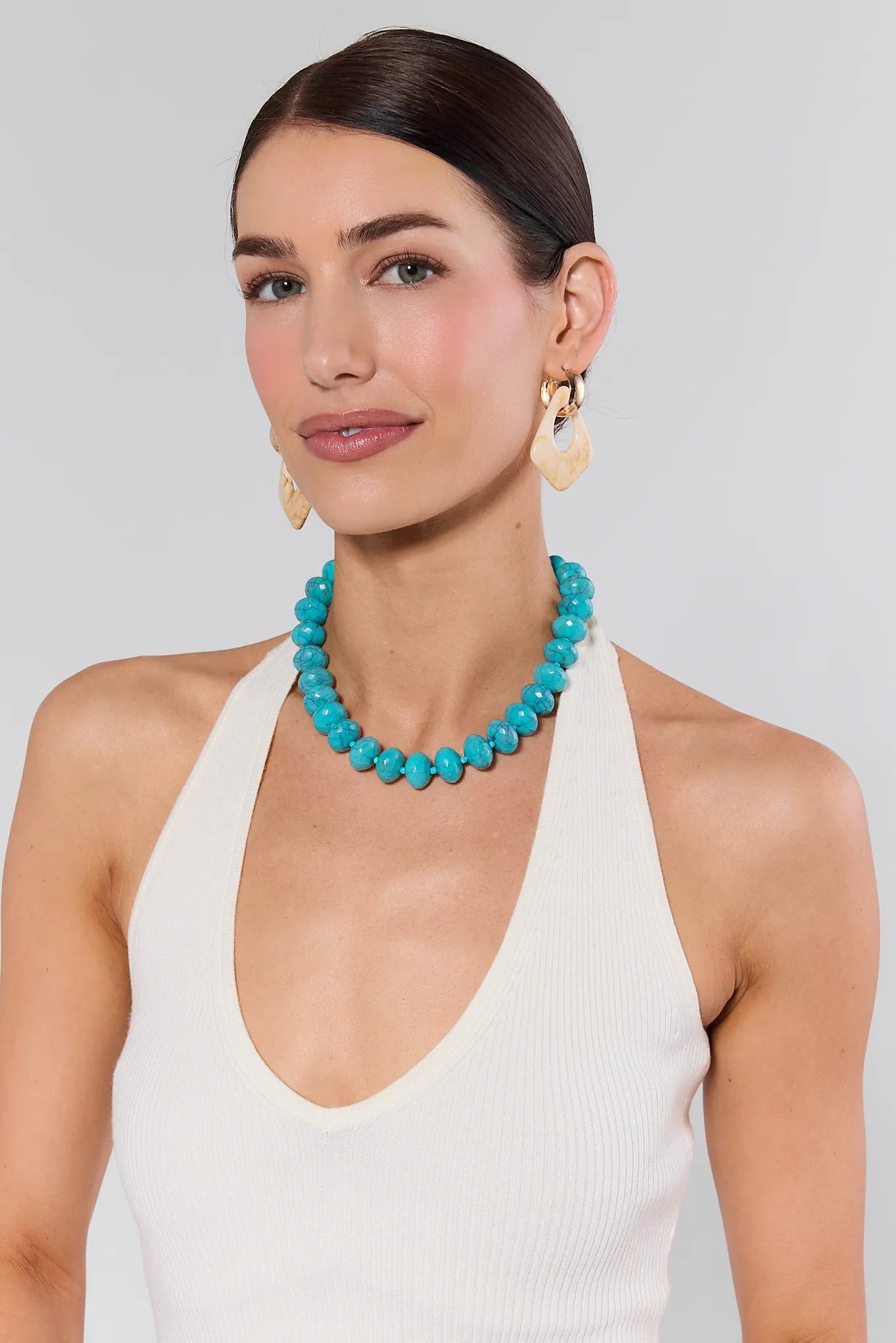 Woman wearing a turquoise necklace and beige earrings against a plain background