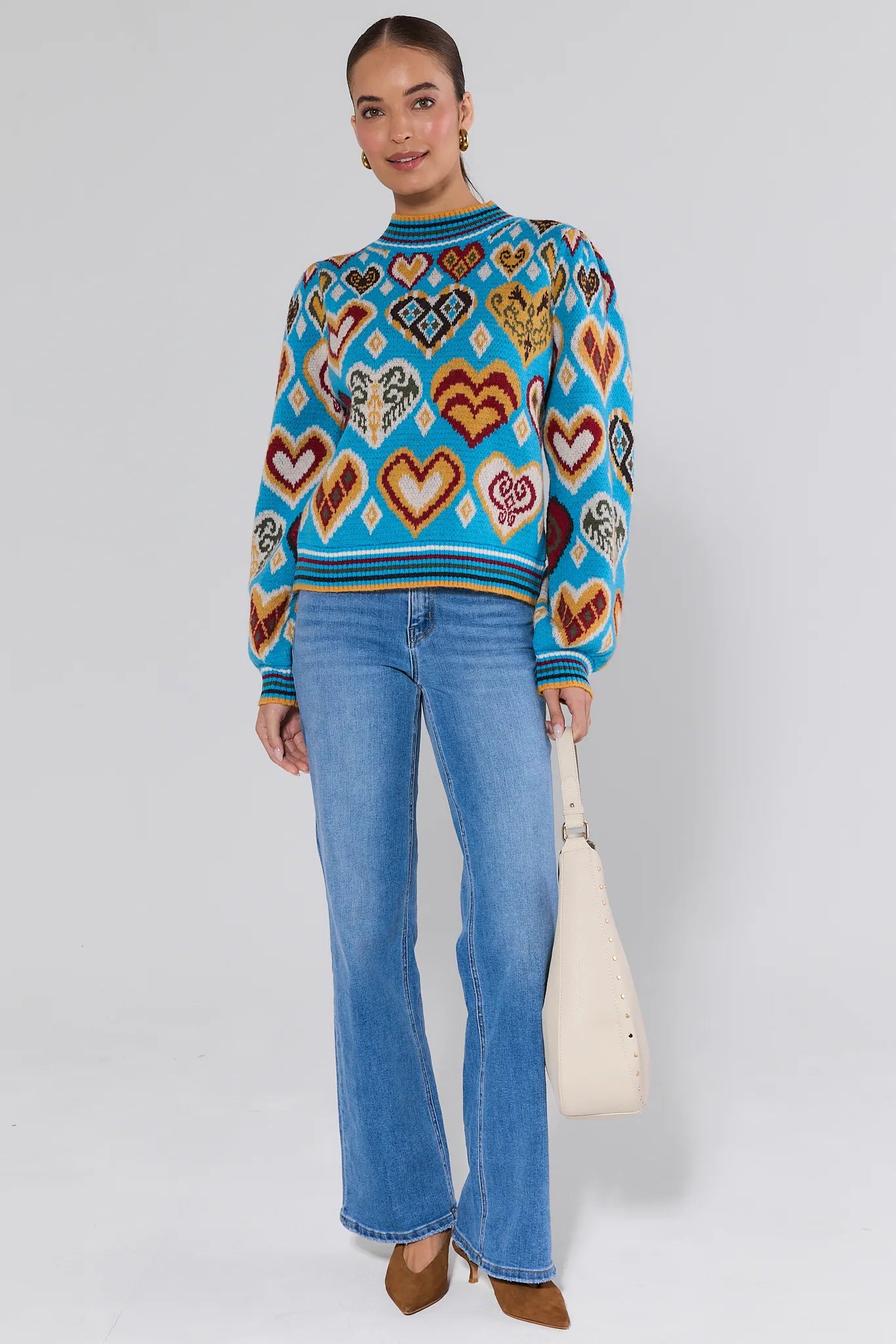 Woman wearing a colorful heart-patterned Cammie Blue Heart Knit Sweater and blue jeans against a plain background