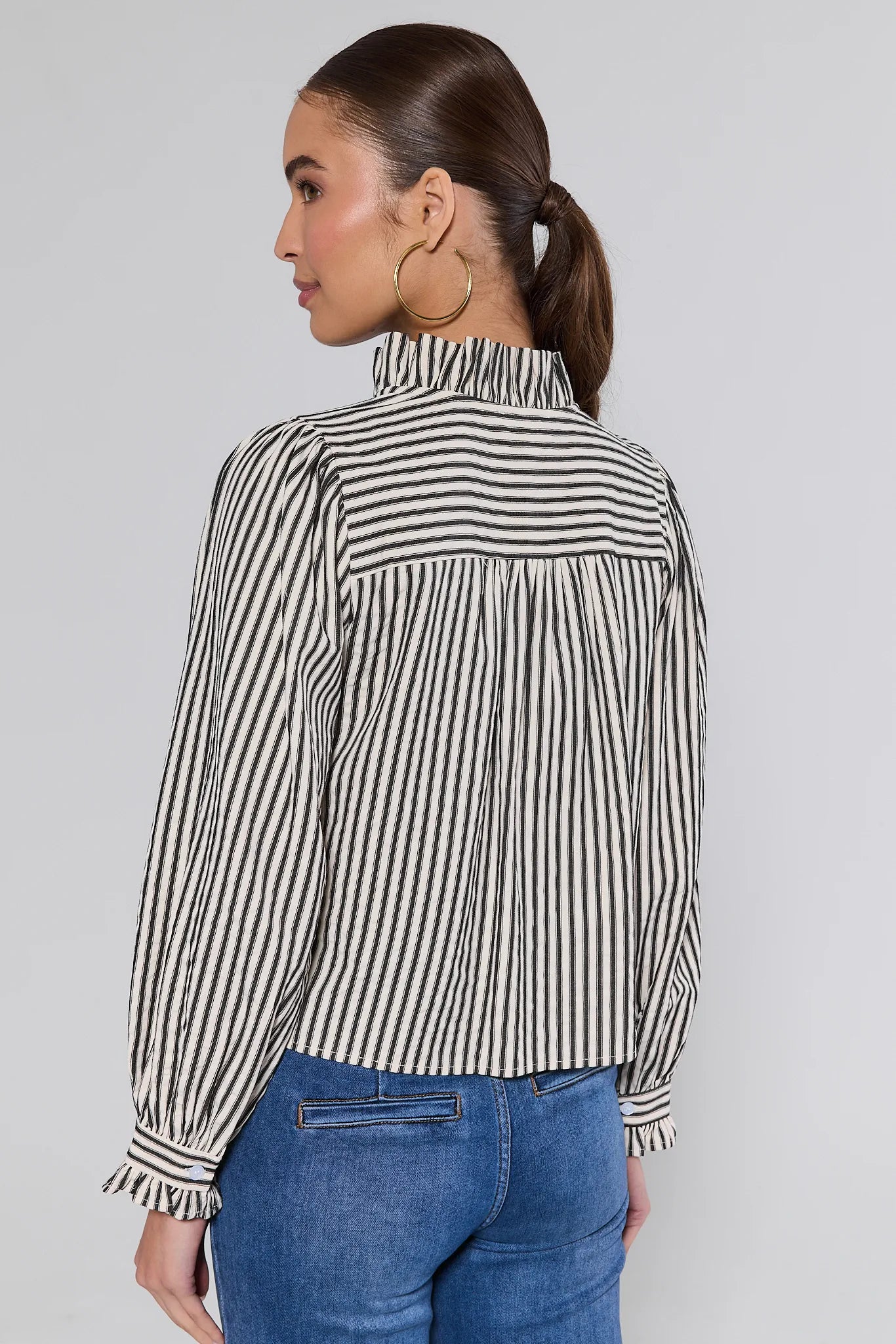 Woman wearing a black and white Jacie Striped Ruffle Long Sleeve Top with blue jeans on a gray background