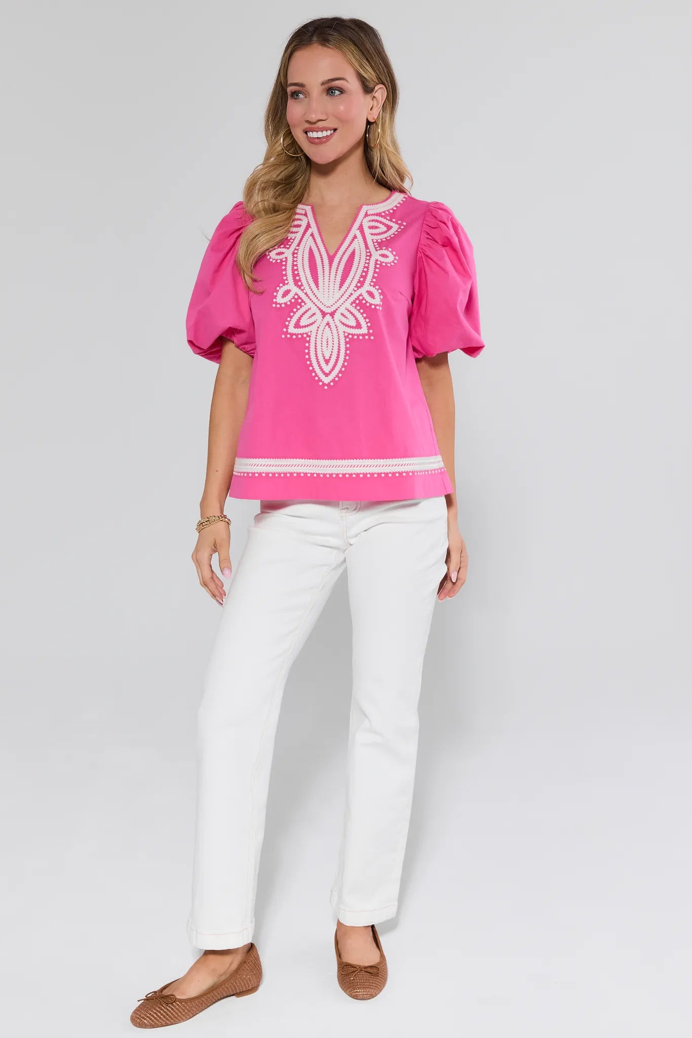 Woman wearing a pink blouse with white embroidery and white pants on a gray background