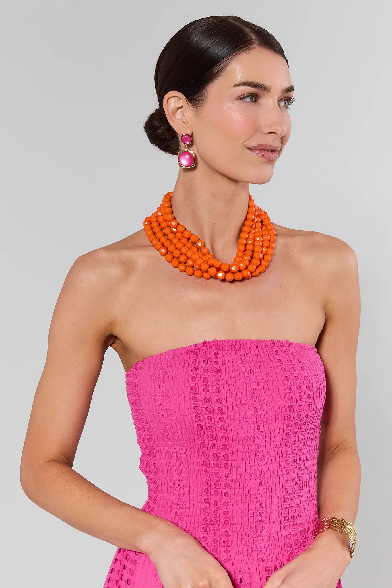 Woman wearing a bright pink strapless dress with an orange necklace on a gray background