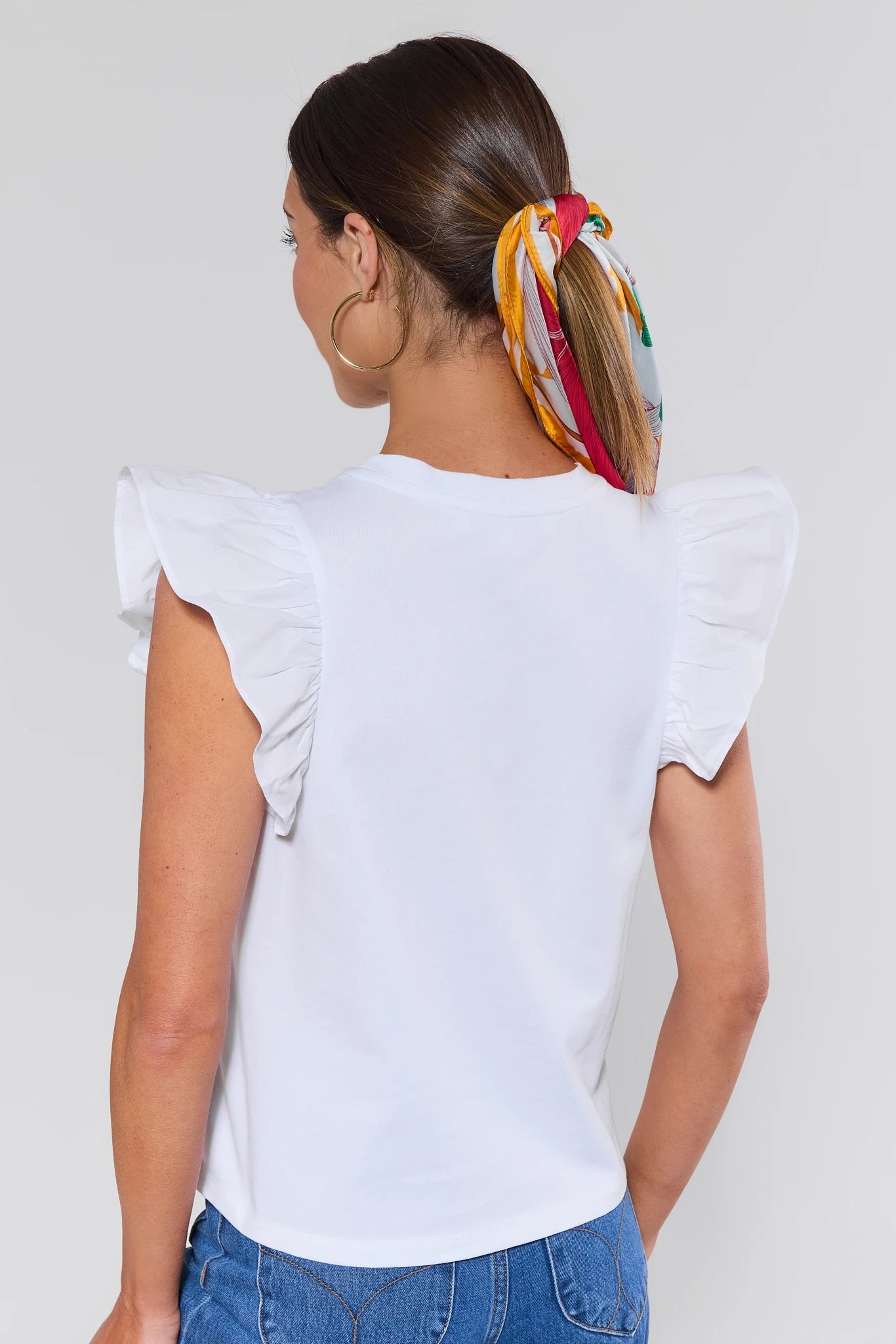 Woman wearing a white top with ruffled sleeves and blue jeans, with a colorful headscarf on her head against a plain background.