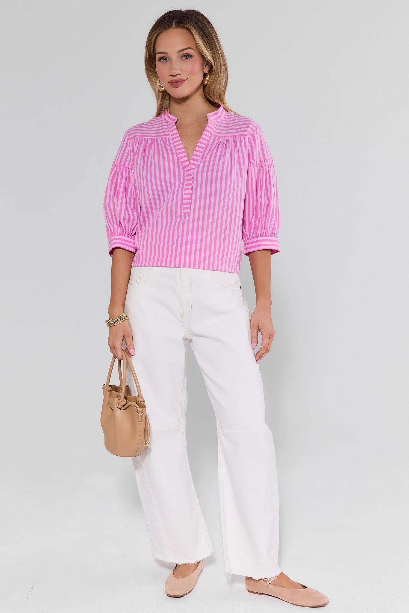 Woman wearing a pink striped shirt and white pants, holding a beige handbag on a light gray background