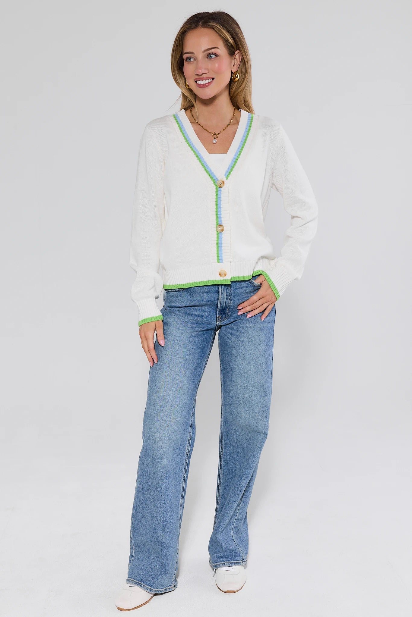 Woman wearing a white cardigan with green trim and blue jeans on a white background
