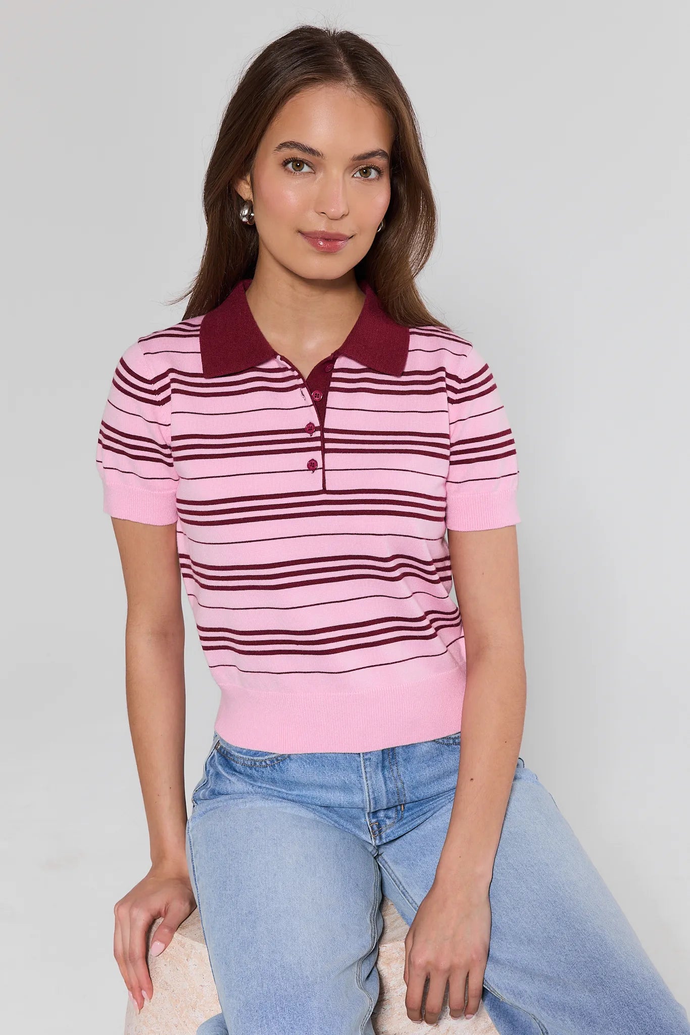 Woman wearing a pink and maroon striped polo shirt with blue jeans on a plain background
