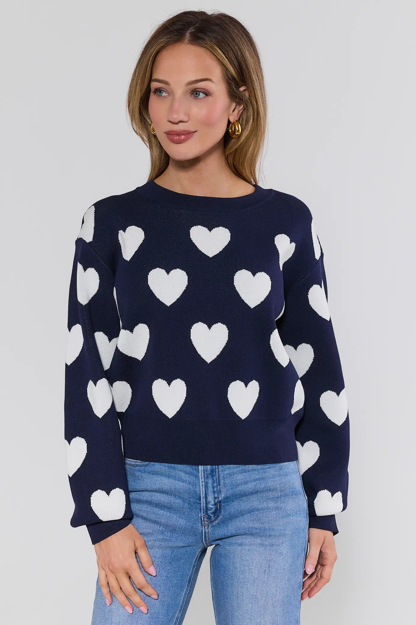 Woman wearing a navy sweater with white heart patterns and blue jeans on a plain background