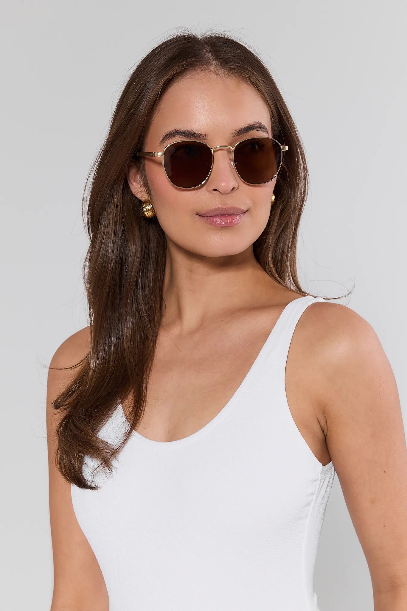 Woman wearing round sunglasses and a white tank top against a plain background