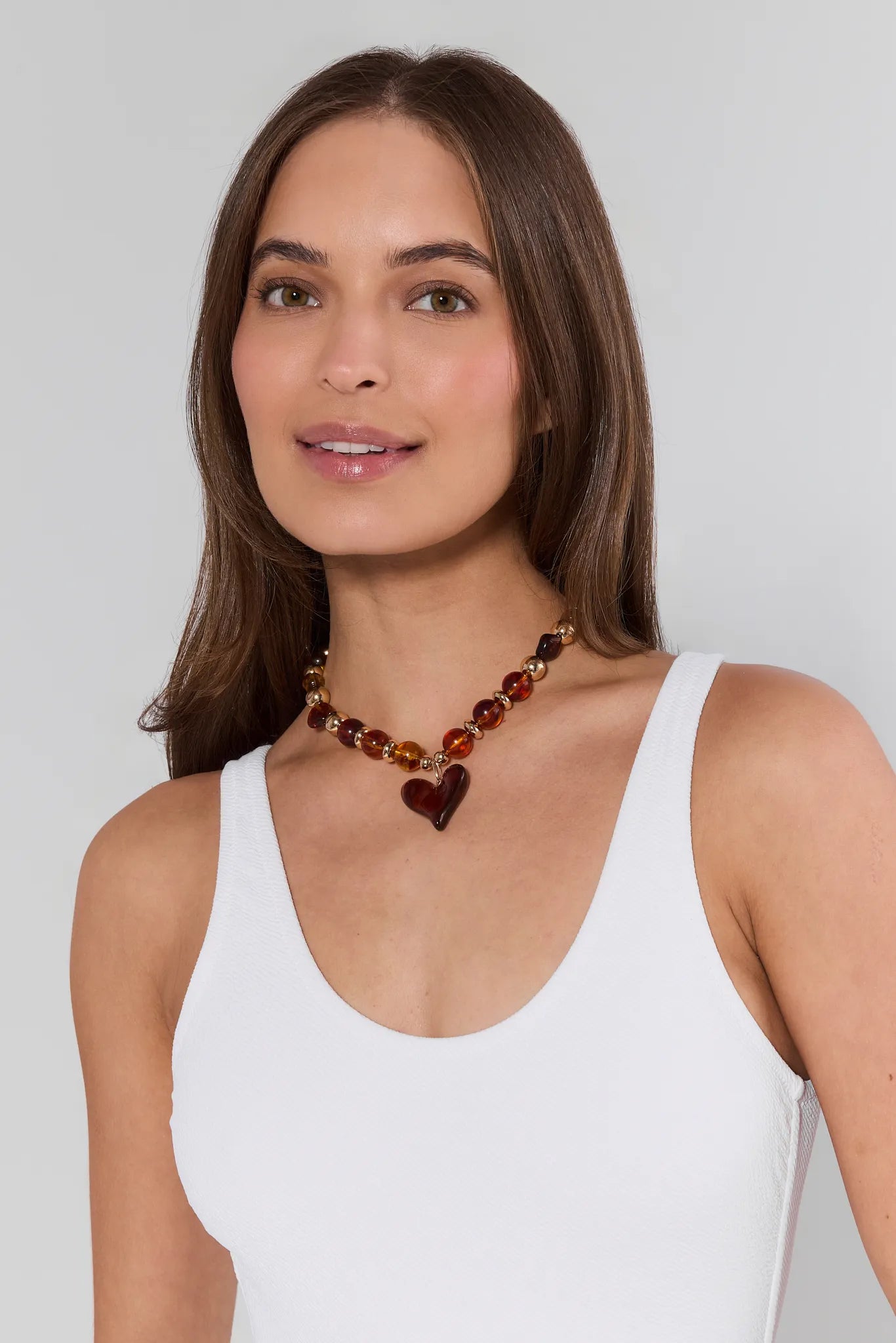 Woman wearing a white tank top and a necklace with heart-shaped beads on a plain background