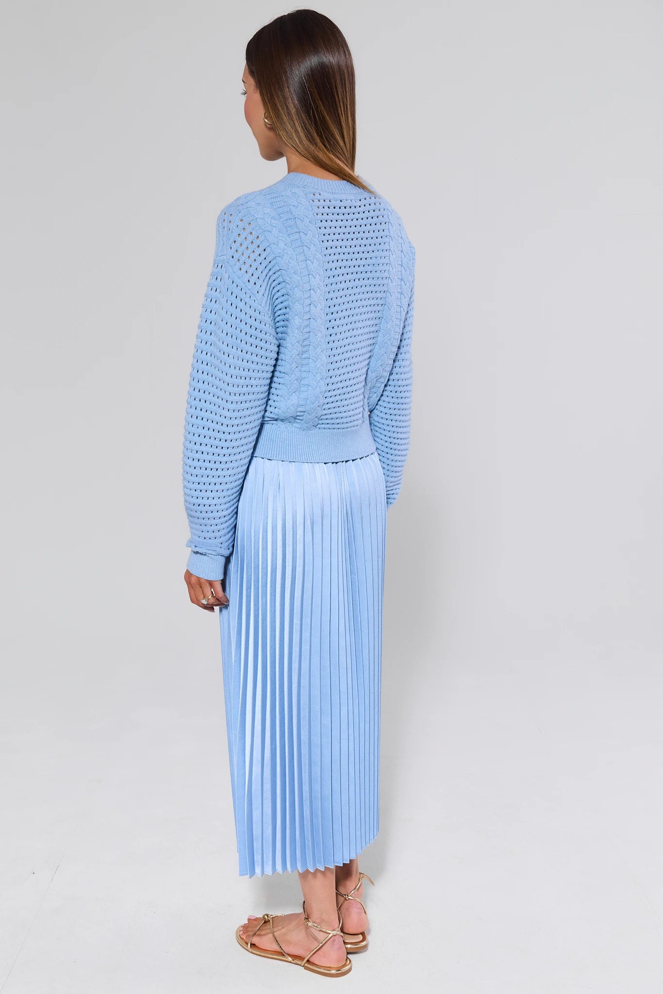 Woman wearing a light blue knitted sweater and pleated skirt against a white background