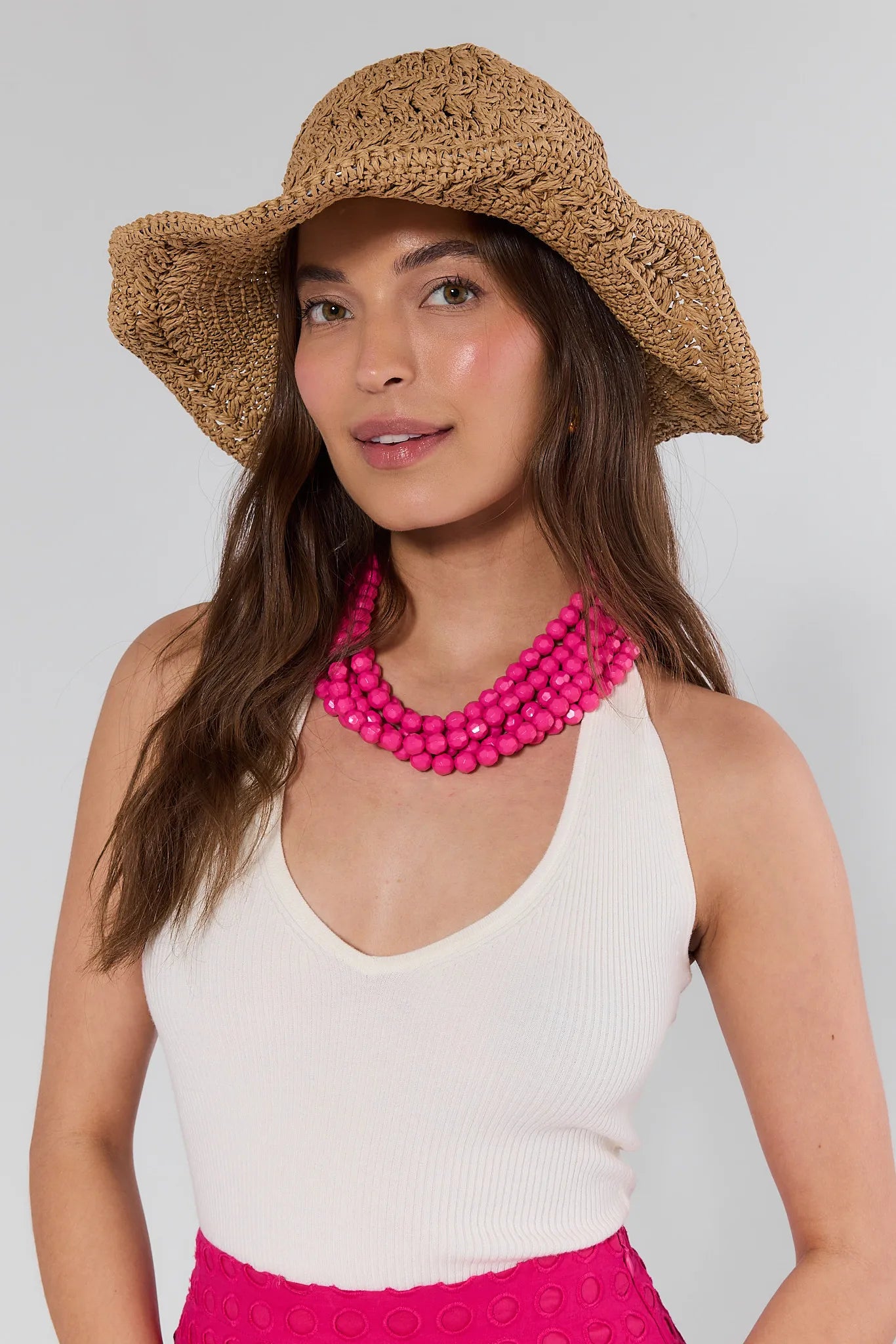 Woman wearing a straw hat, white top, and pink skirt with a pink necklace on a plain background