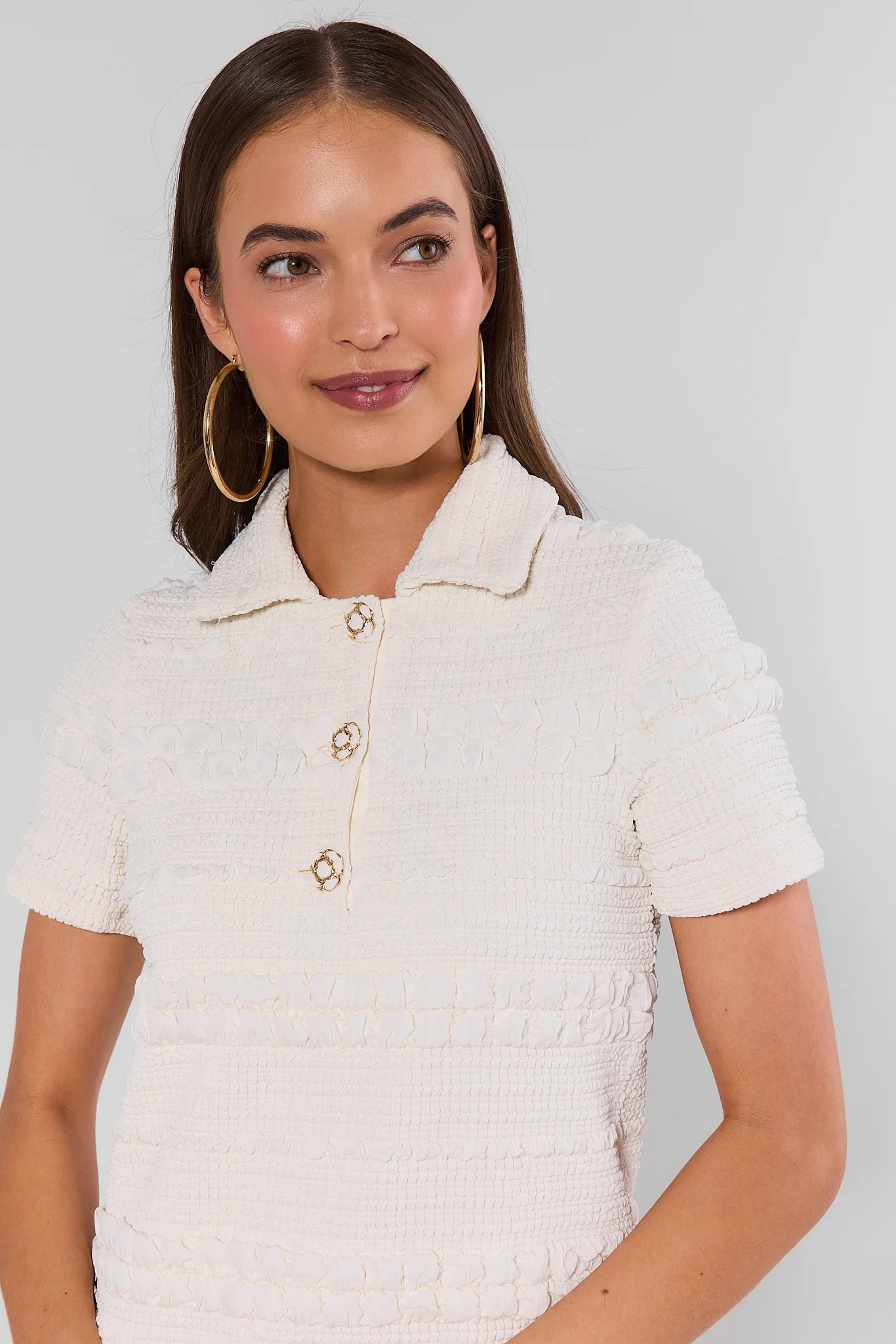 Woman wearing a textured white top with gold buttons against a plain background