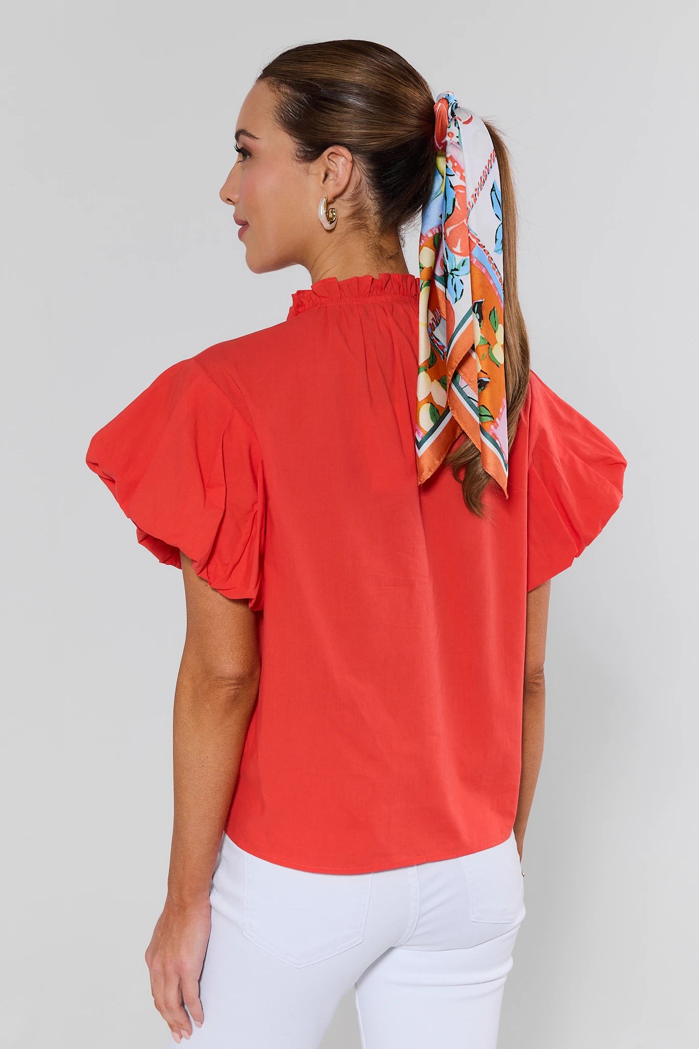 Woman wearing a red blouse with white pants and a colorful scarf on her head against a plain background