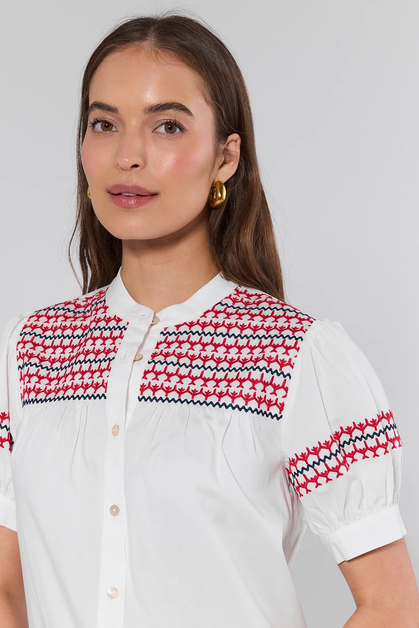 Woman wearing a white shirt with red and black patterned shoulders against a light gray background .