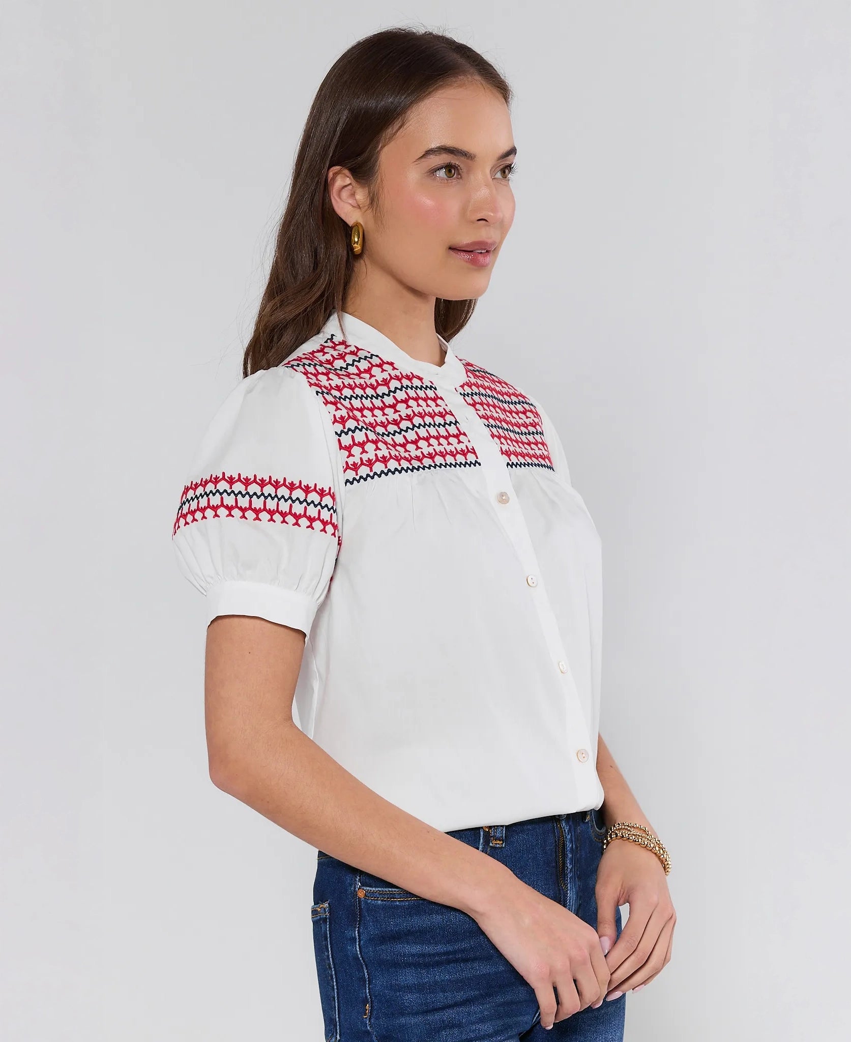 Woman standing at an angle wearing a white blouse with red and black patterned shoulders on a plain background
