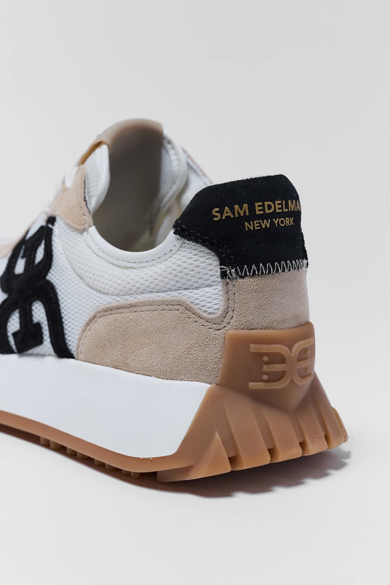 Close-up of a white sneaker with brown sole and black and gold accents on a light gray background.