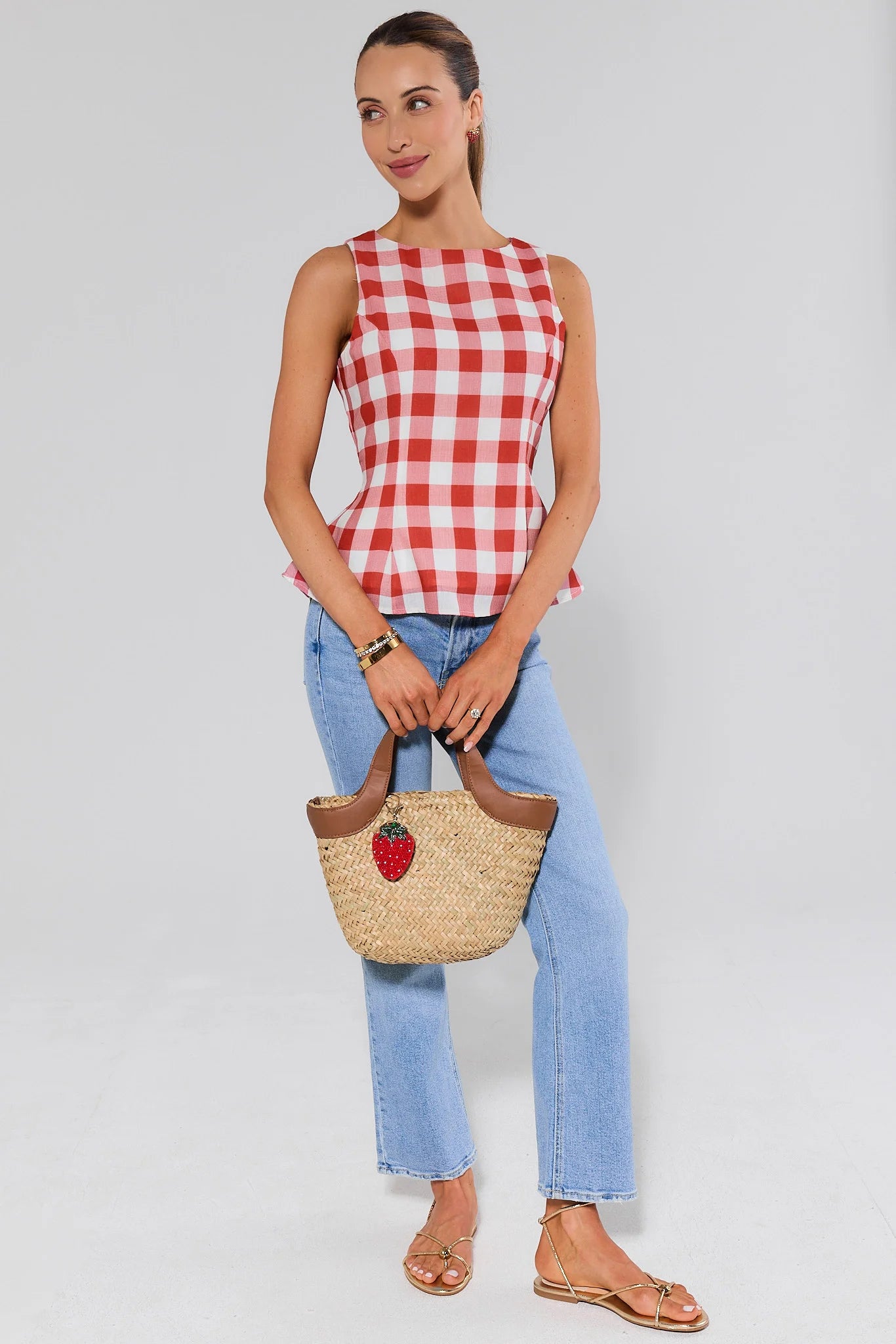 Woman wearing a red and white checkered top and blue jeans, holding a straw bag with a strawberry design.