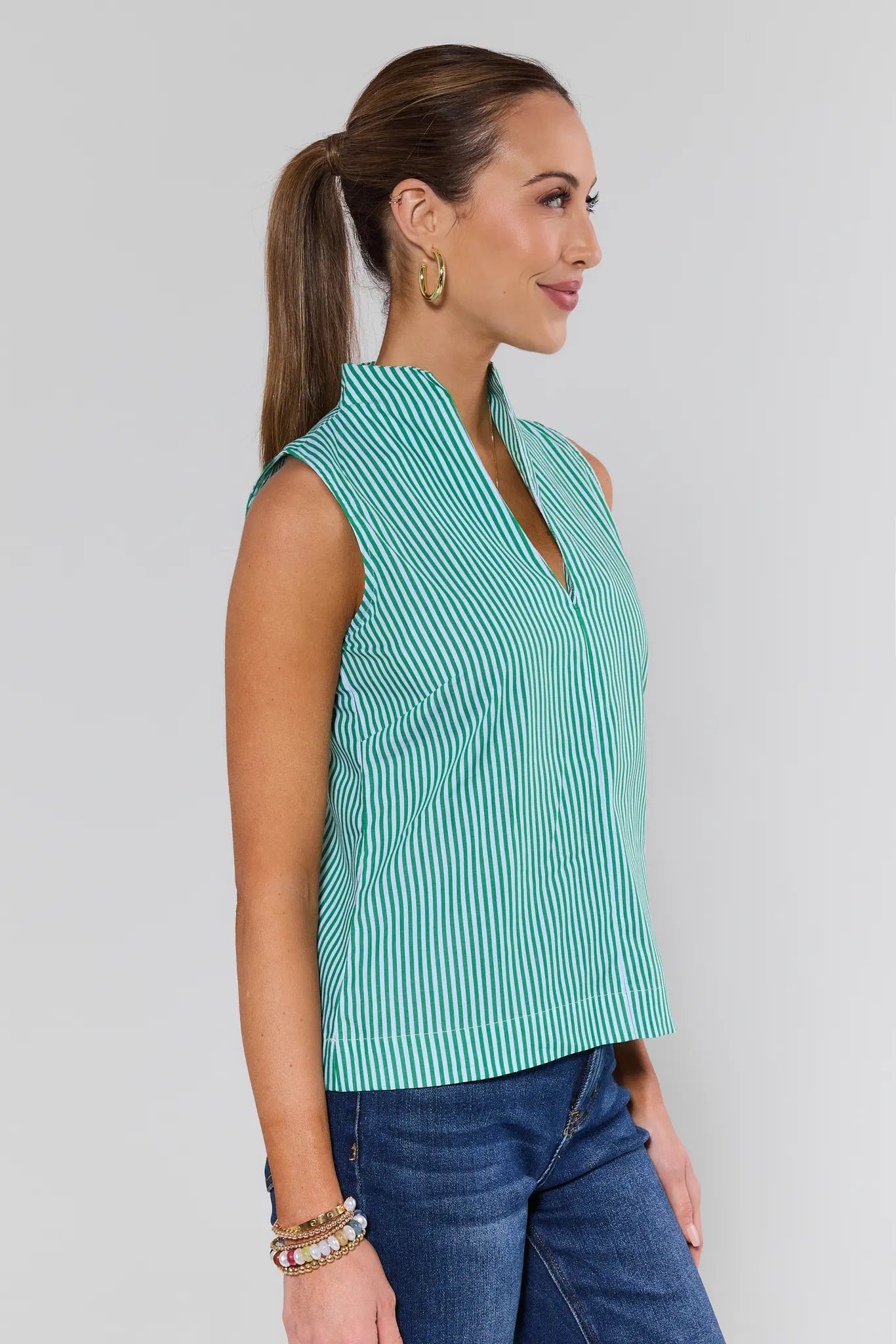 Woman wearing a green and white striped sleeveless top with blue jeans on a plain background
