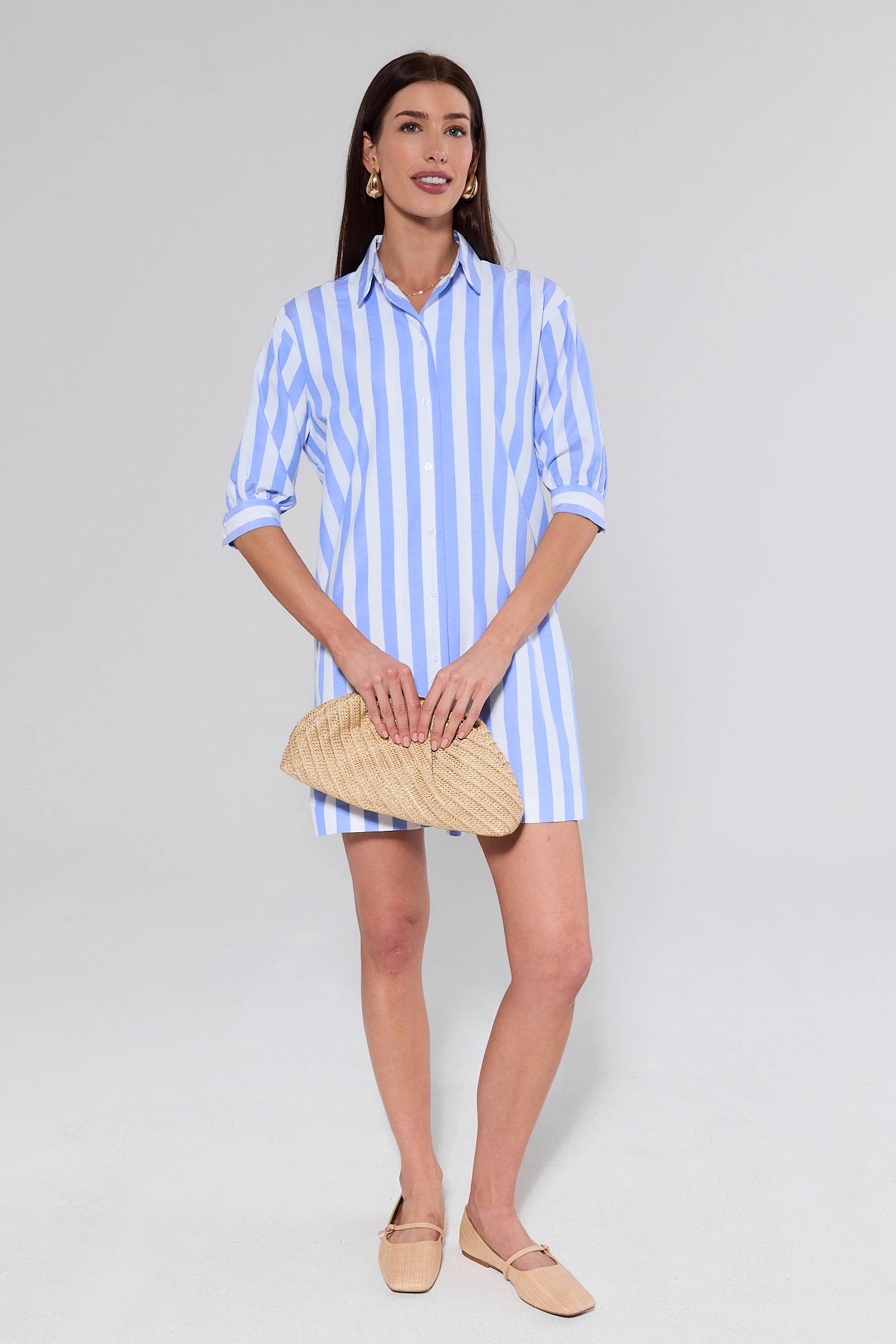 Woman wearing a blue and white striped dress holding a straw bag on a plain background