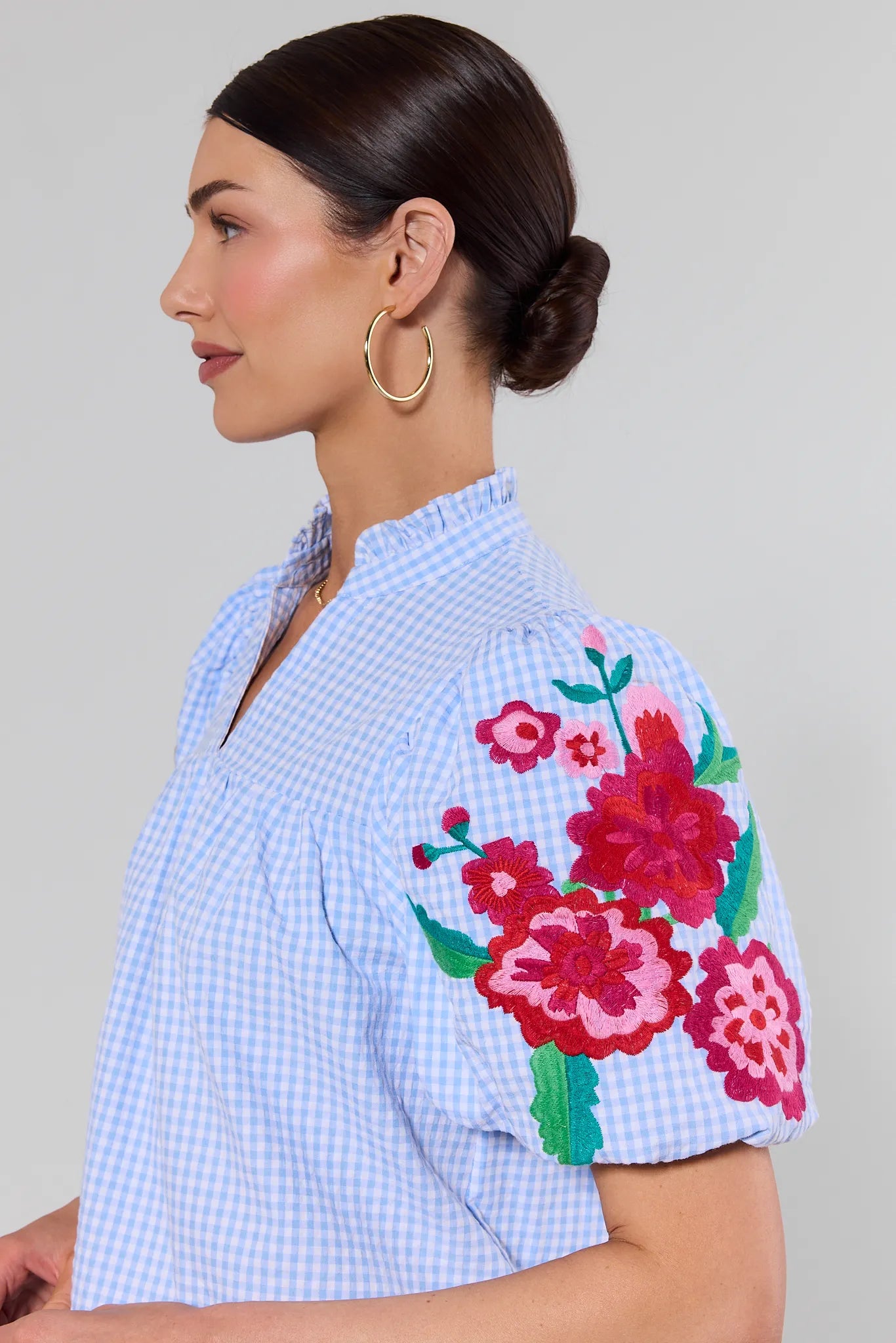 close up Woman wearing a light blue checkered shirt with floral embroidery on sleeves against a gray background