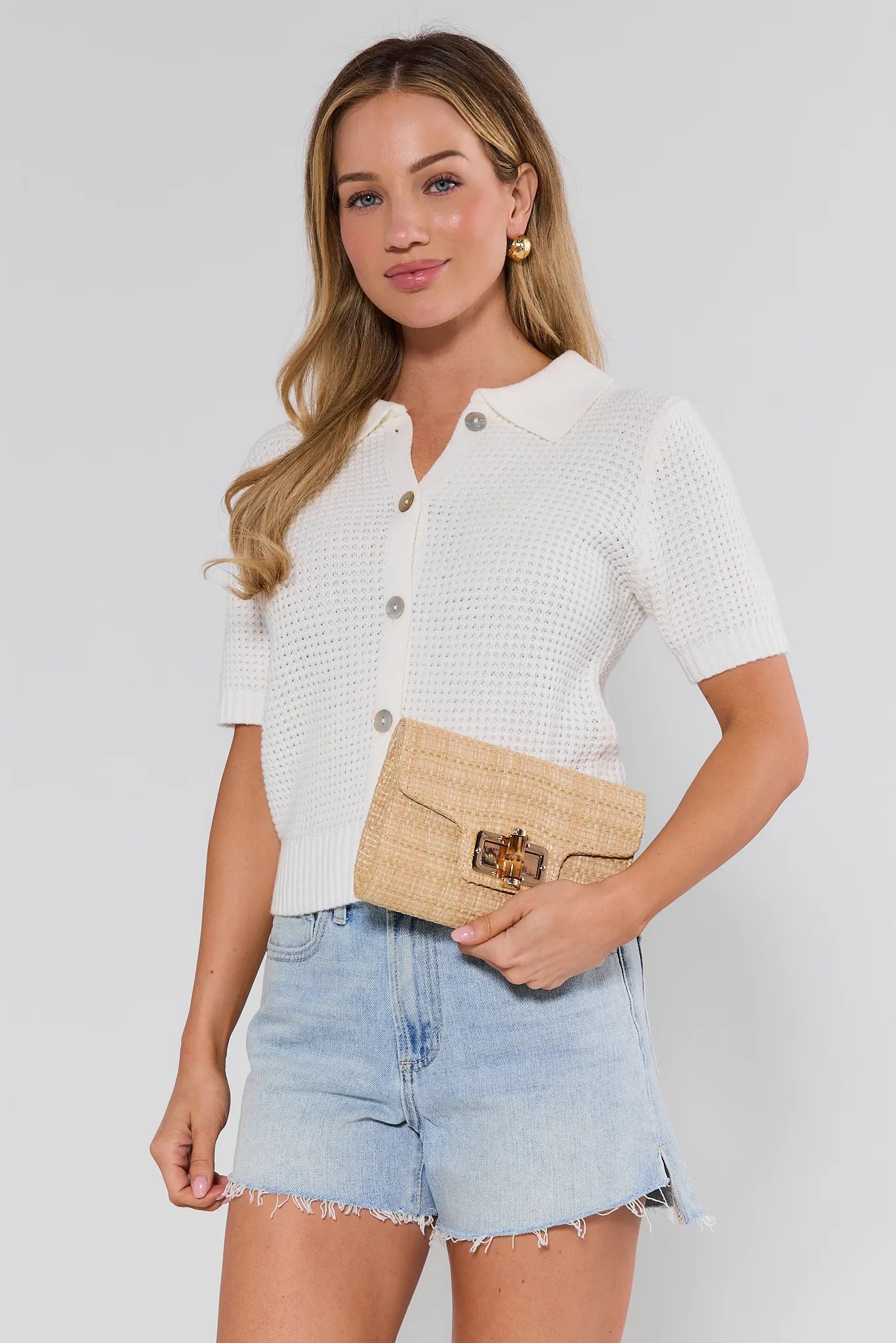 Woman wearing a white button-up shirt and denim shorts, holding a beige clutch against a plain background