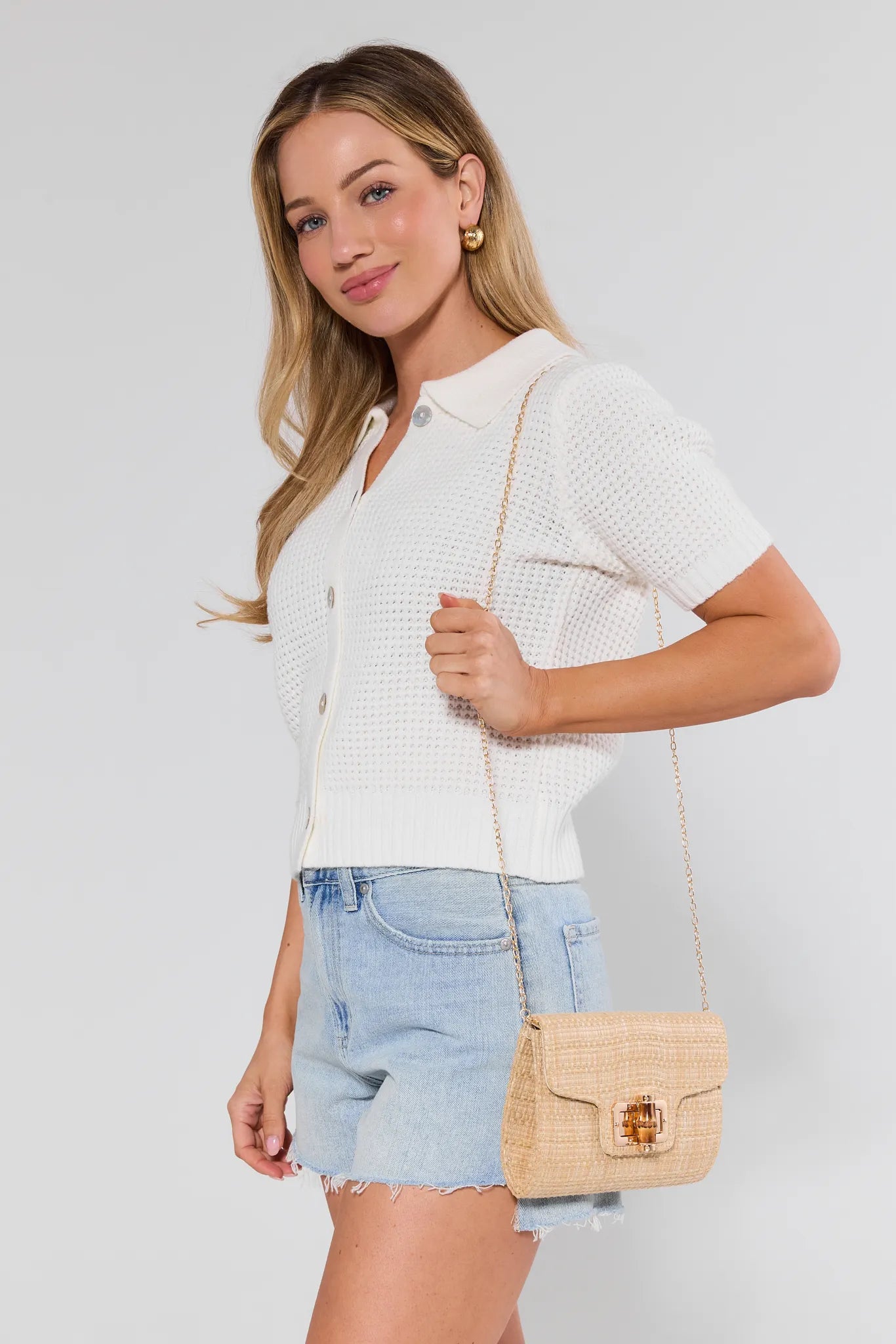 Woman wearing a white textured shirt and denim shorts, holding a beige handbag against a plain background