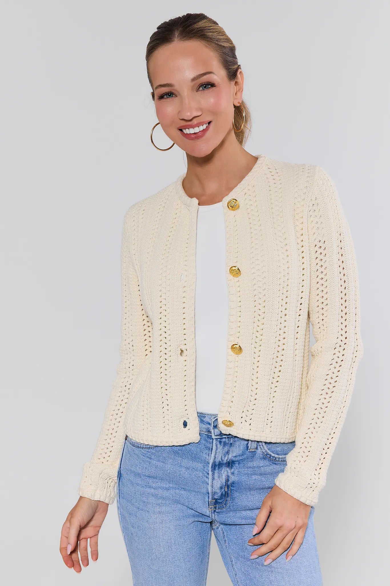 Woman wearing a cream knitted cardigan with gold buttons on a plain background