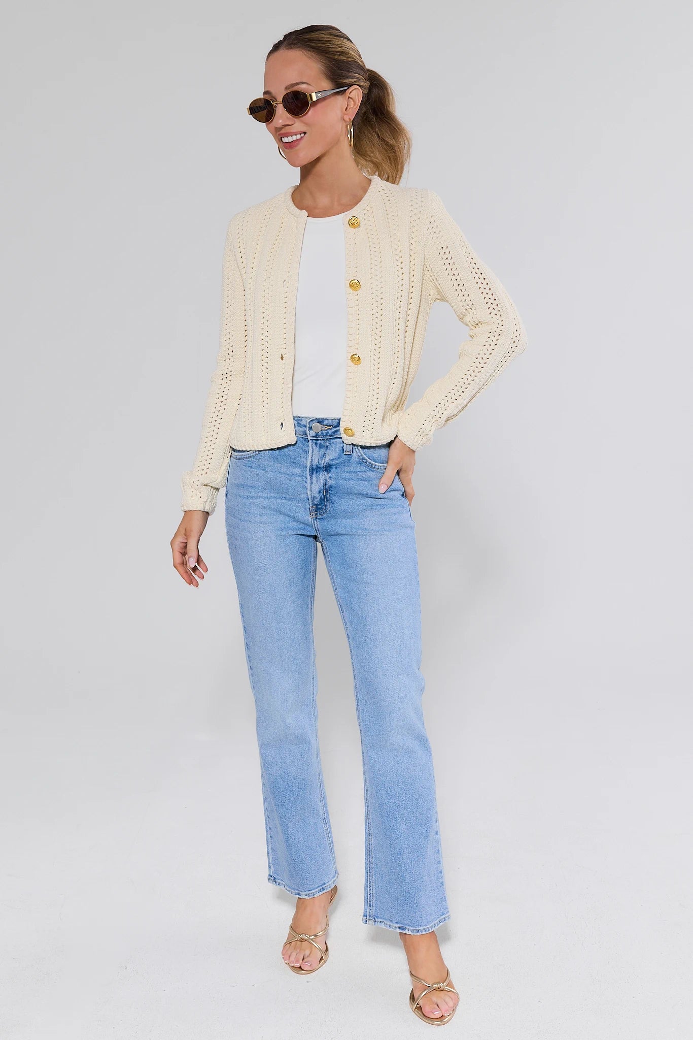 Woman wearing a beige cardigan, white shirt, and light blue jeans on a plain background