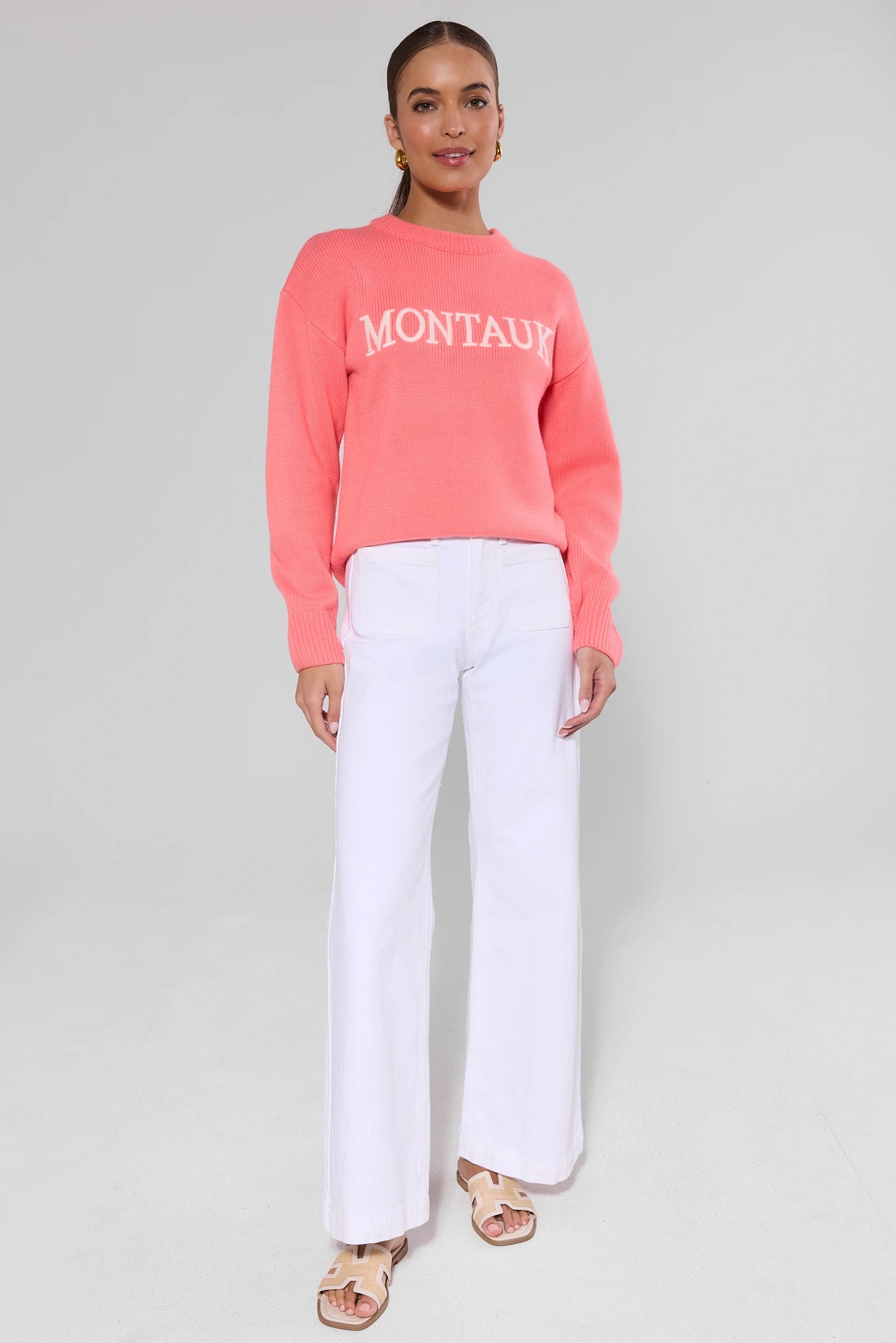 Woman wearing a coral sweatshirt with 'Montauk' text and white pants on a gray background