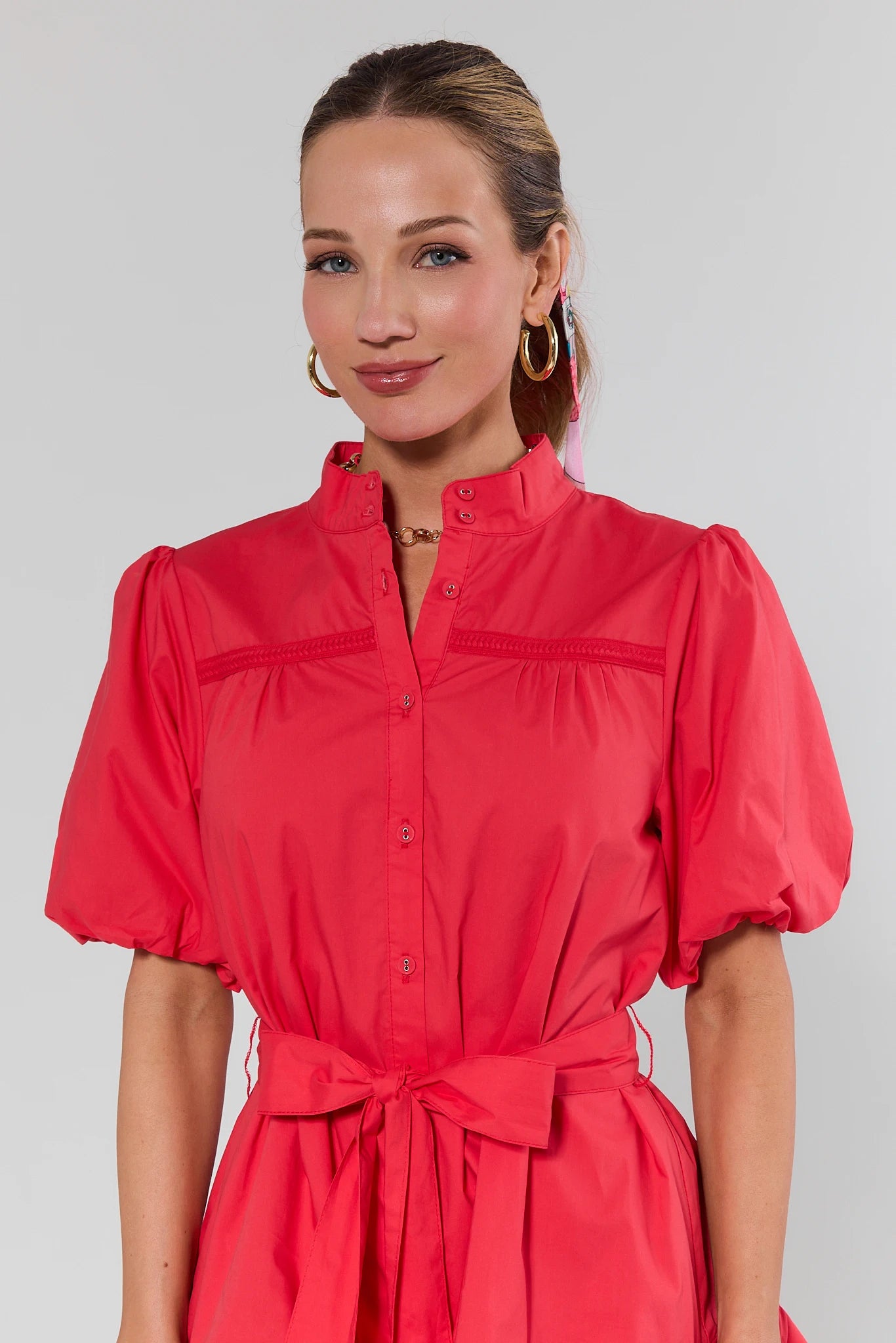 Model wearing Jane Red Puff Sleeve Short Dress with braided trim, button-front, puffed sleeves, self-tie belt, and a-line silhouette, accessorized with gold jewelry.