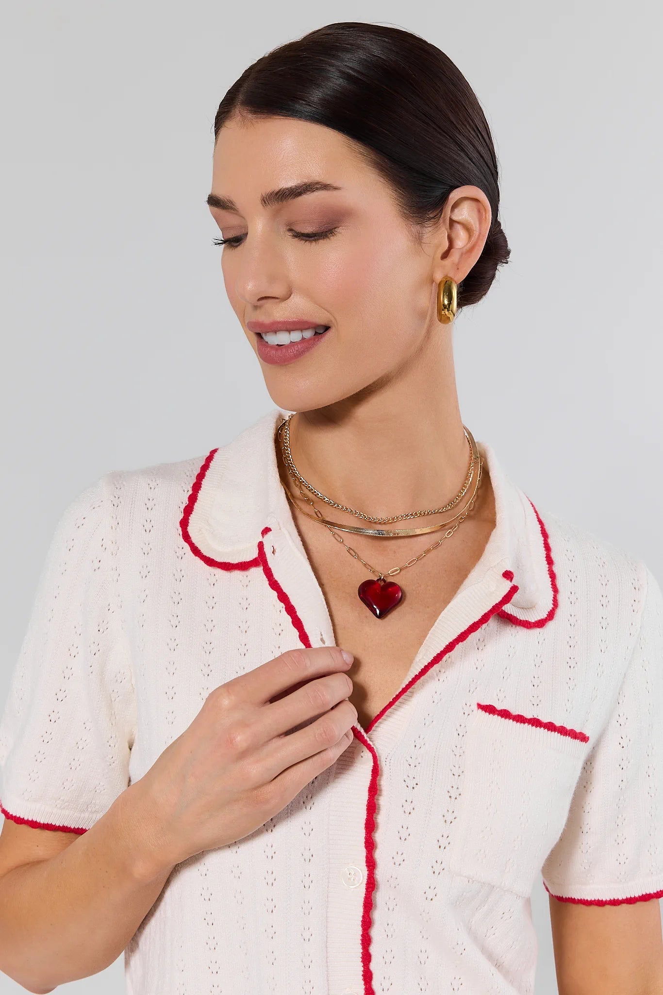 Woman wearing a white outfit with red trim and gold jewelry against a light gray background