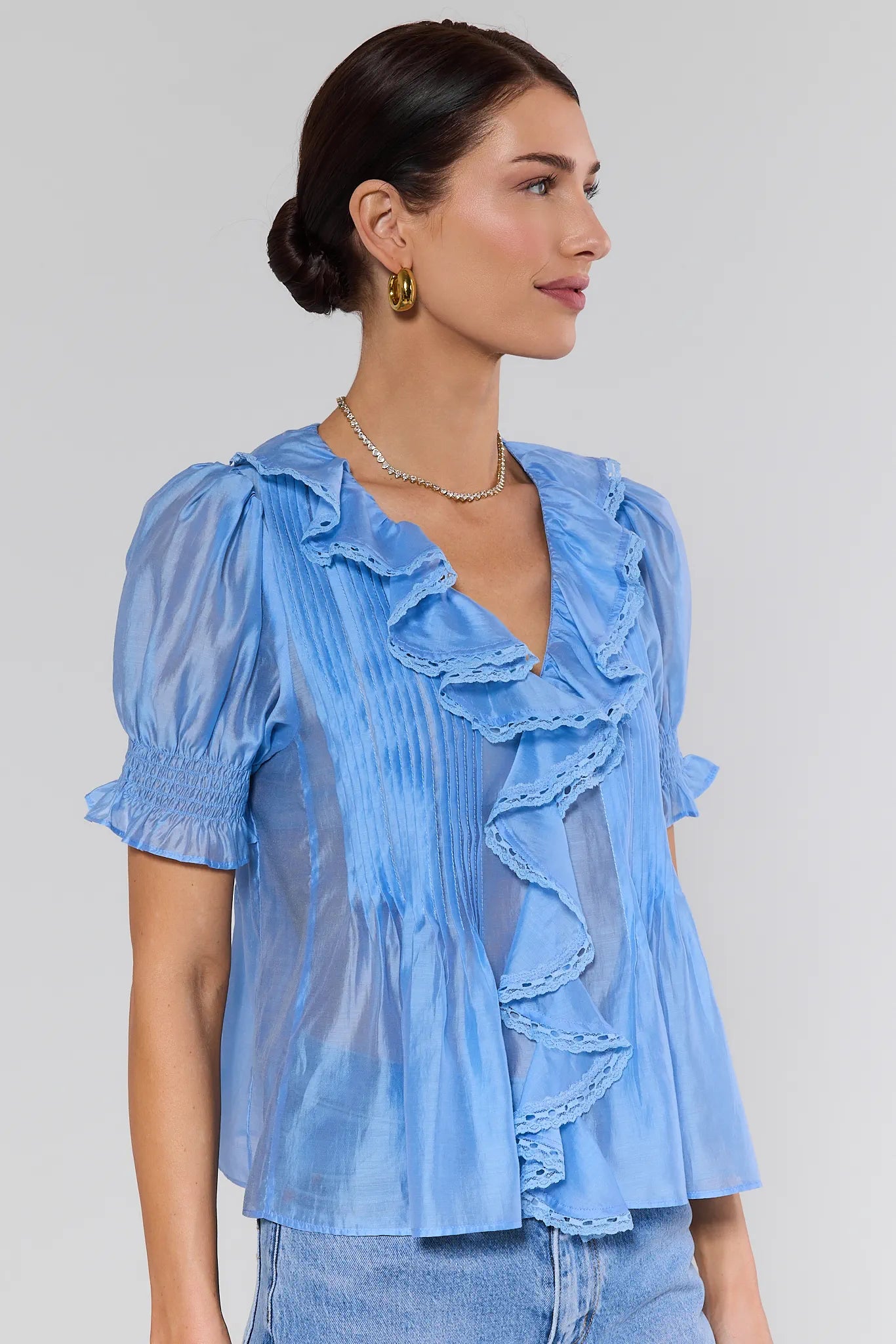 Woman wearing a light blue blouse with ruffled details with gold jewelry on a plain background