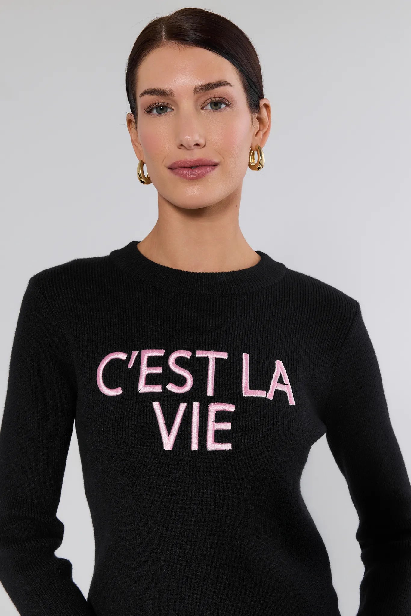 Woman wearing a black sweater with 'C'EST LA VIE' text in pink on a plain background