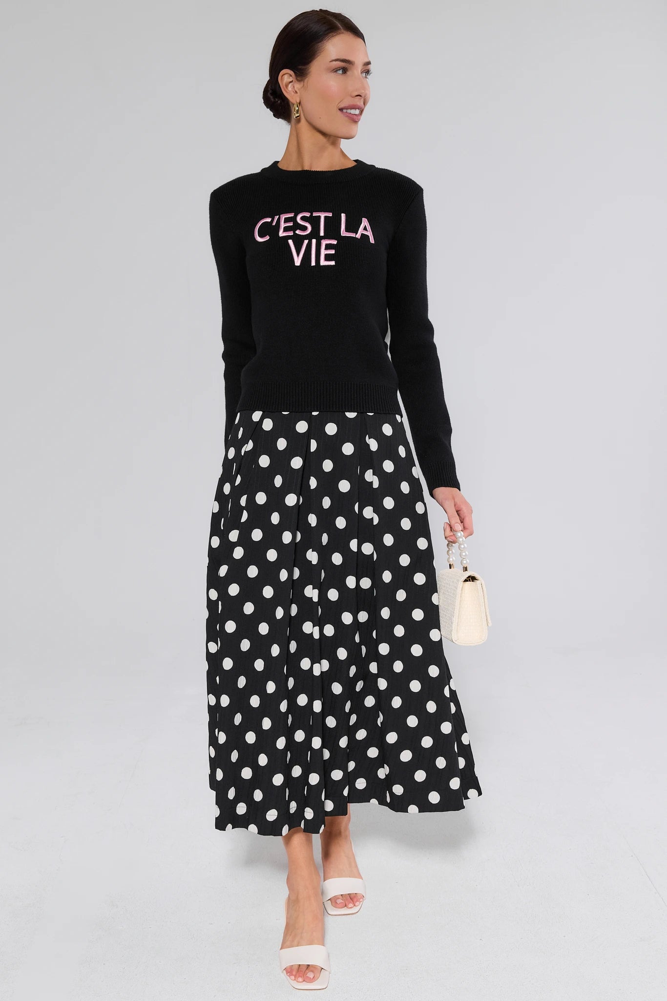 Woman wearing a black sweater with 'C'EST LA VIE' text and a black polka dot skirt on a gray background