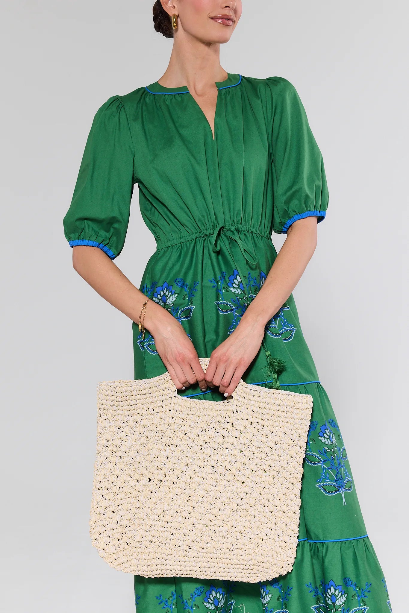 Woman wearing a green dress with floral embroidery, holding a woven beige bag against a plain background