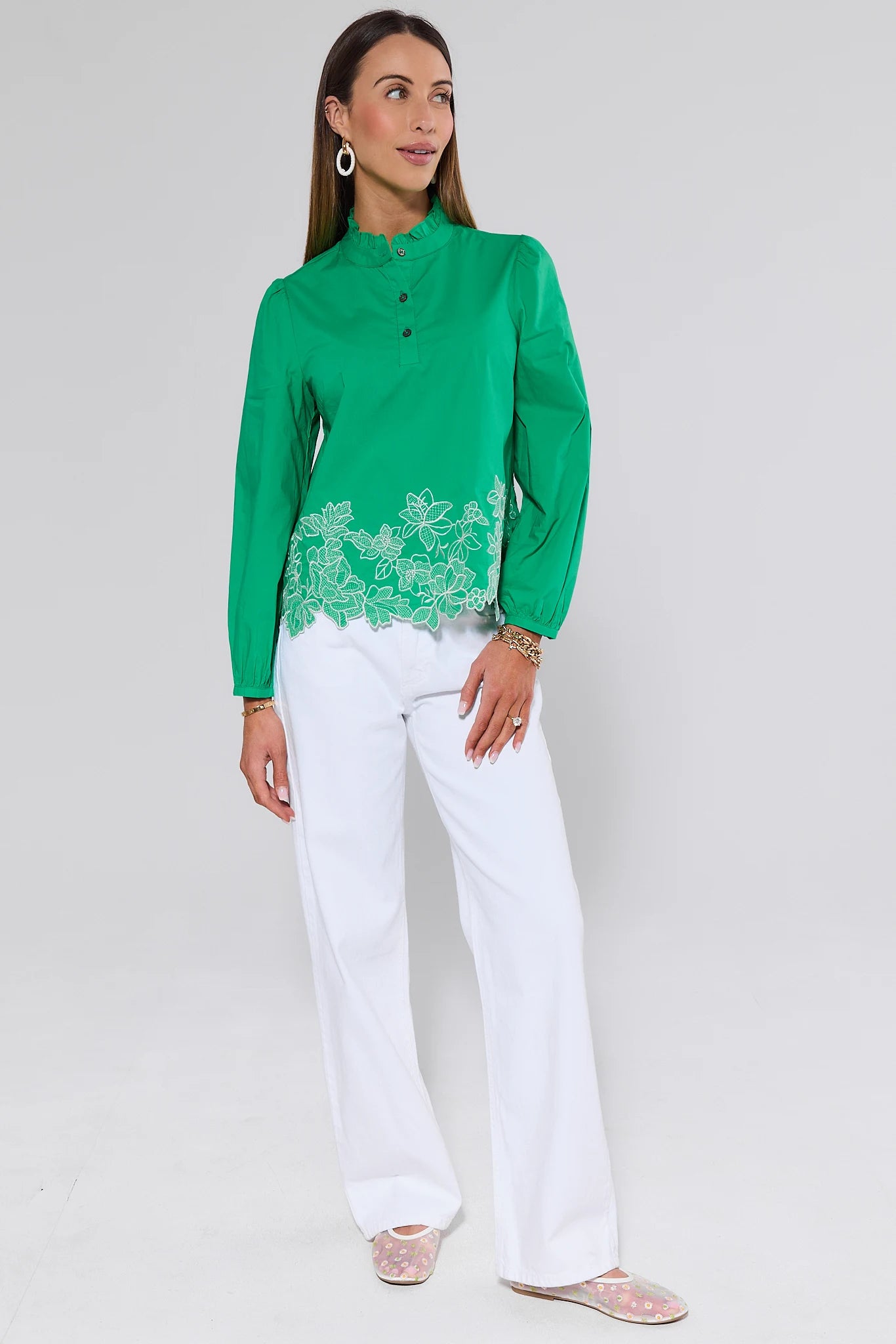 Woman wearing a green blouse with floral embroidery and white pants on a light gray background