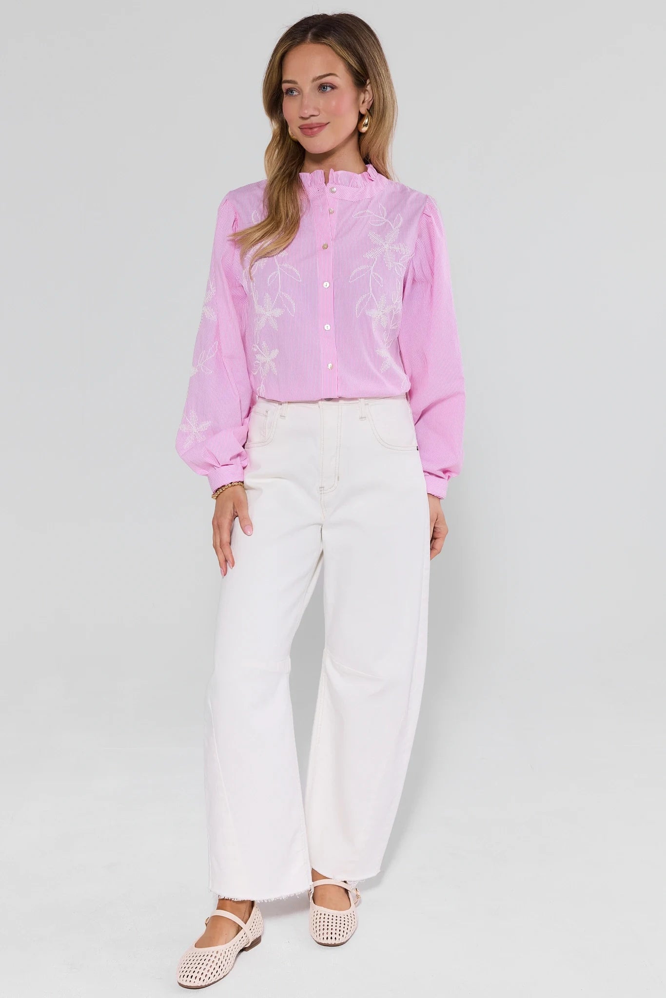 Woman wearing a pink blouse with lace details and white pants on a light gray background