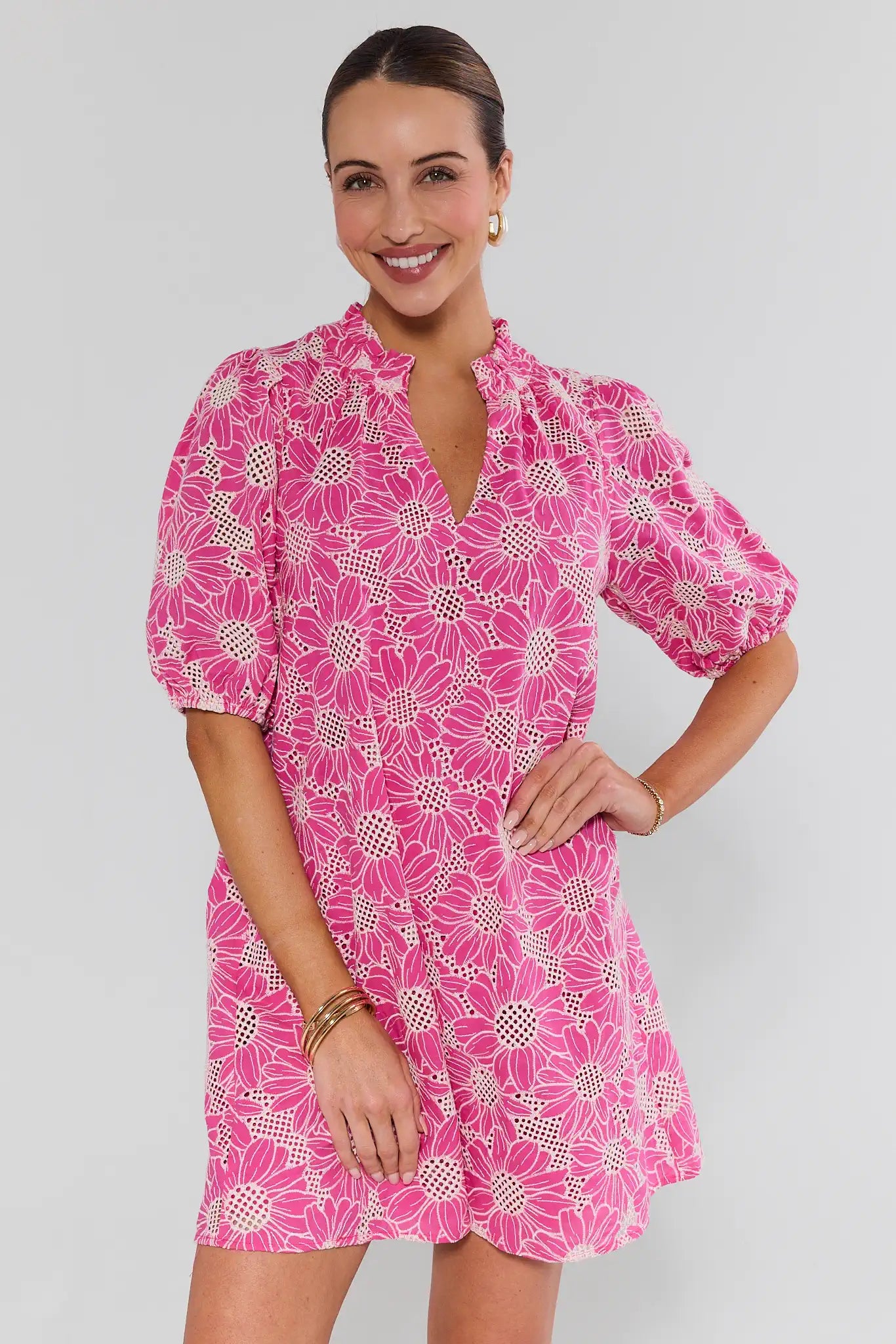 Woman models Bianca Pink Floral Eyelet Short Dress with daisy motif, ruffled collar, V-neck, puff sleeves, and gold accessories, showcasing a chic, comfortable style.