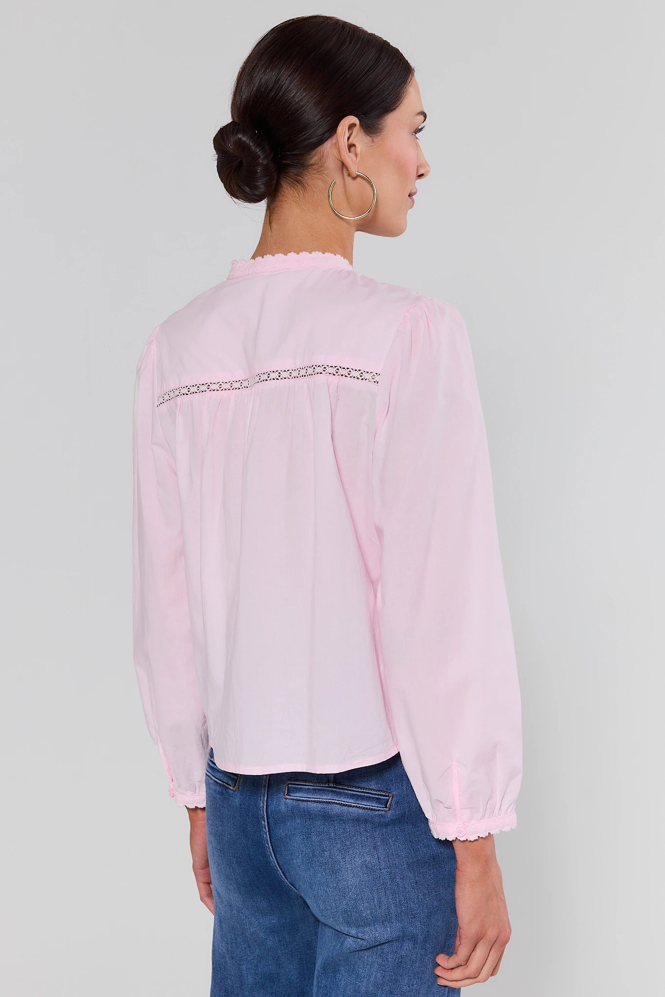 Woman standing wearing a light pink blouse with lace details on a plain background