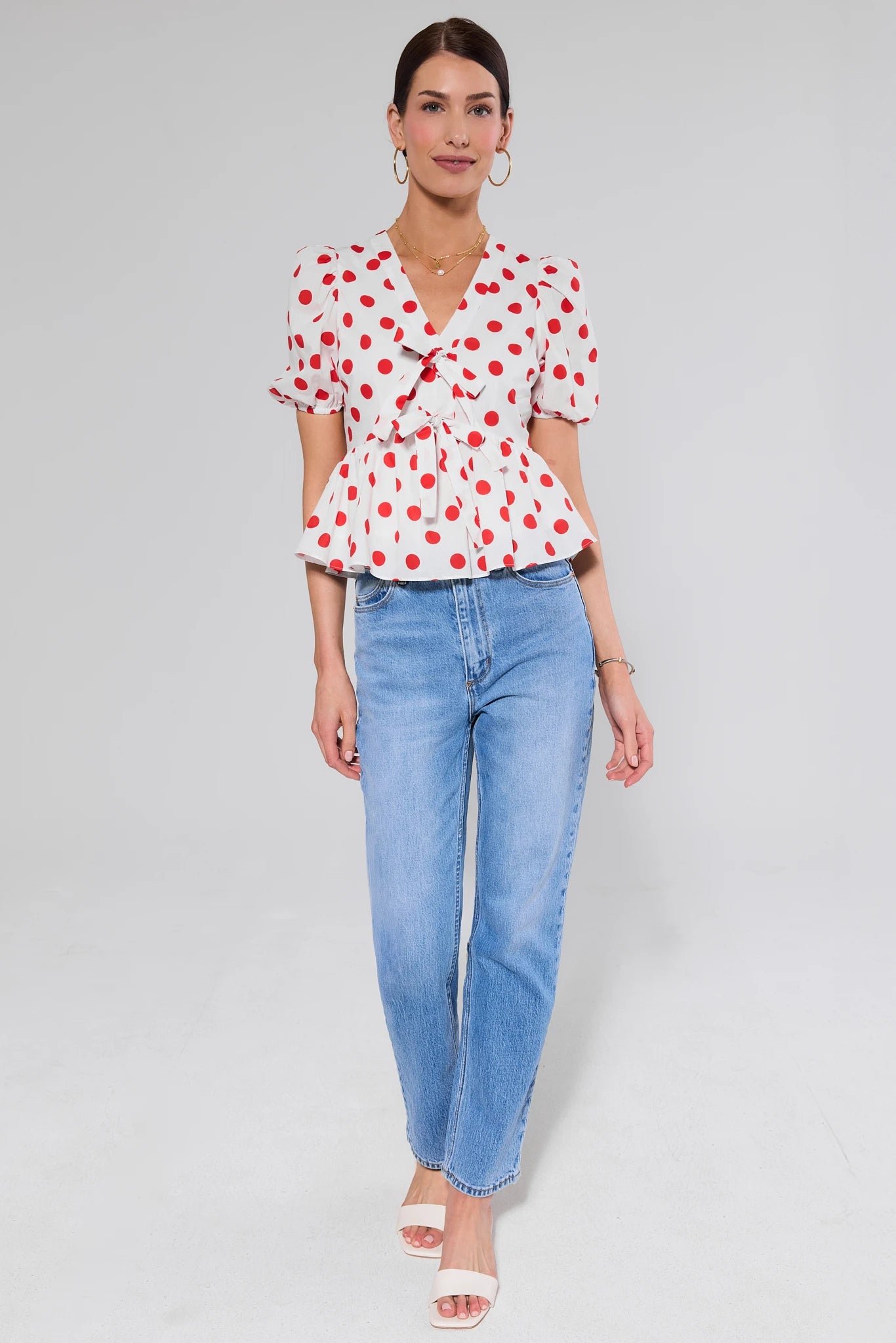 Woman wearing a white blouse with red polka dots and blue jeans on a light gray background