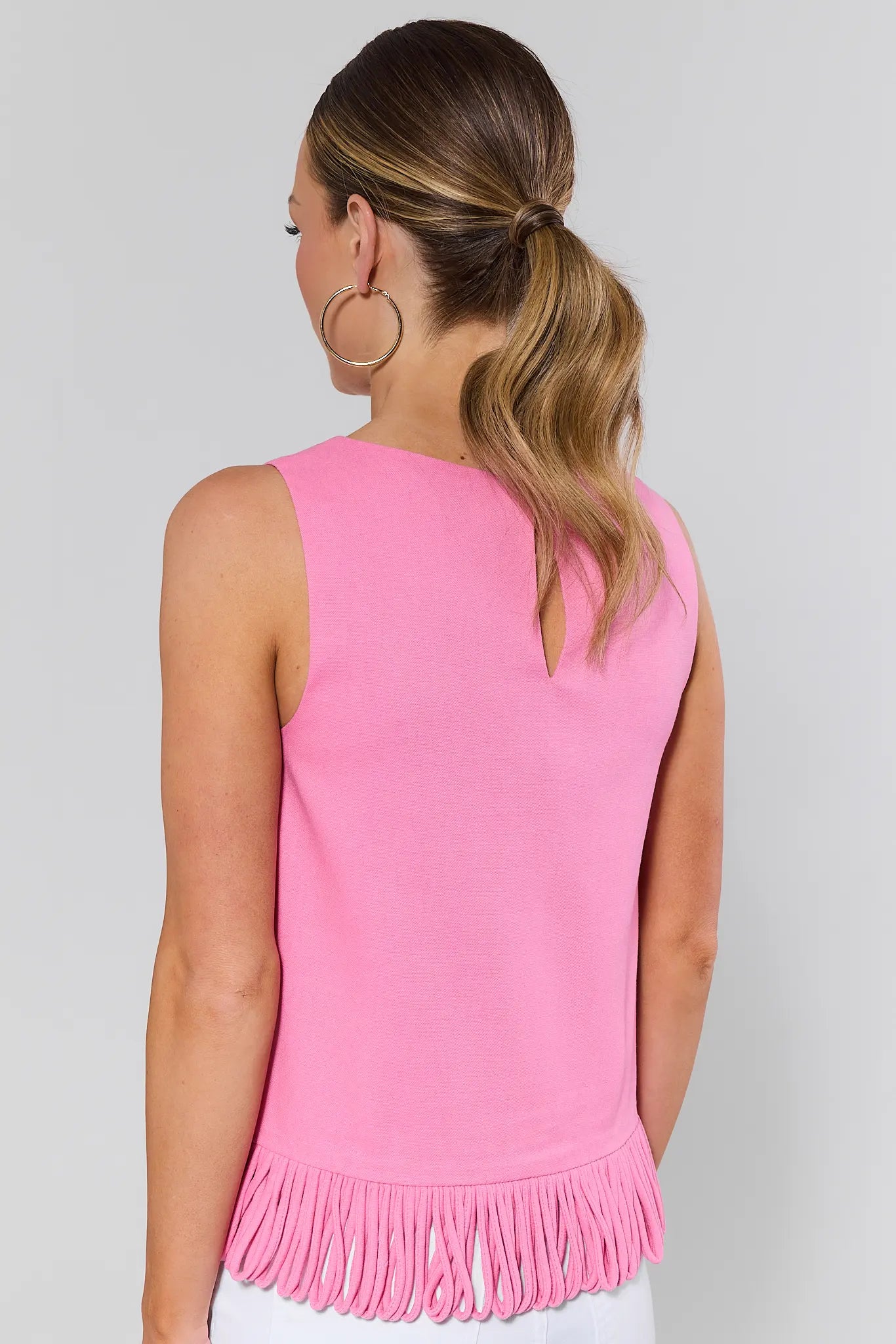 Woman standing wearing a pink sleeveless top with fringe details on a plain background
