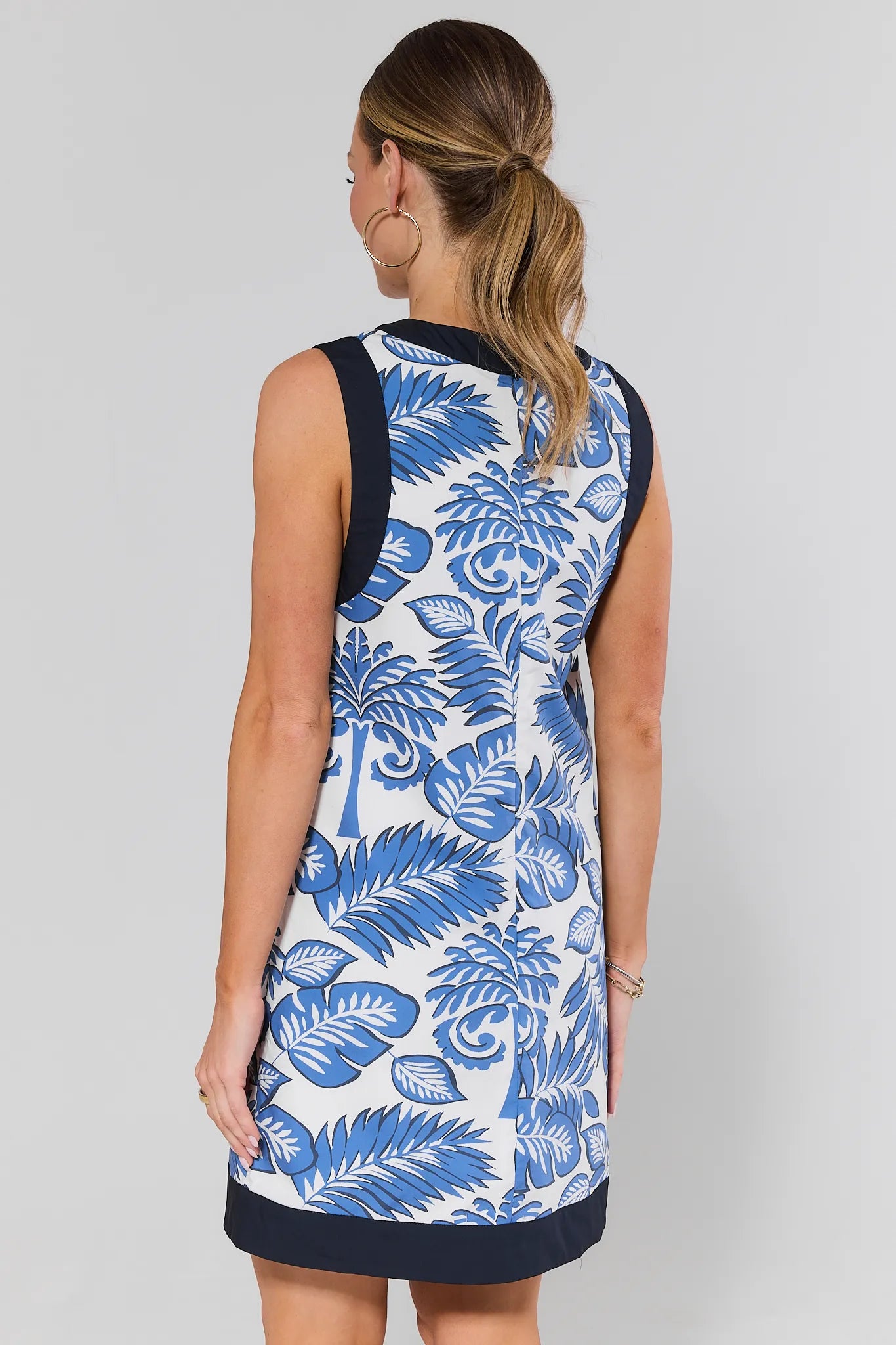 Woman standing wearing a sleeveless dress with blue floral pattern on a plain background