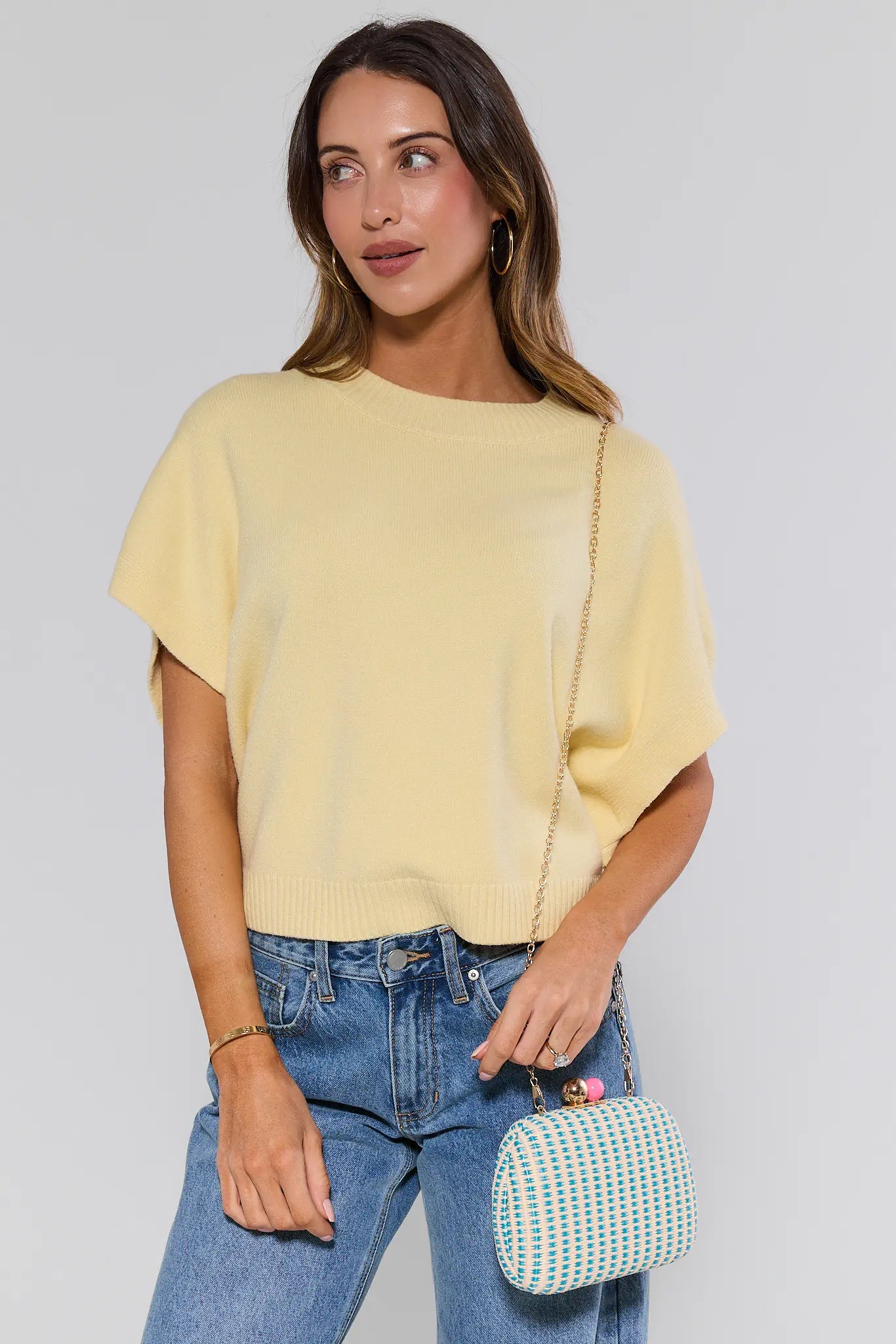 Woman wearing a yellow top and blue jeans, holding a green and white patterned handbag against a plain background