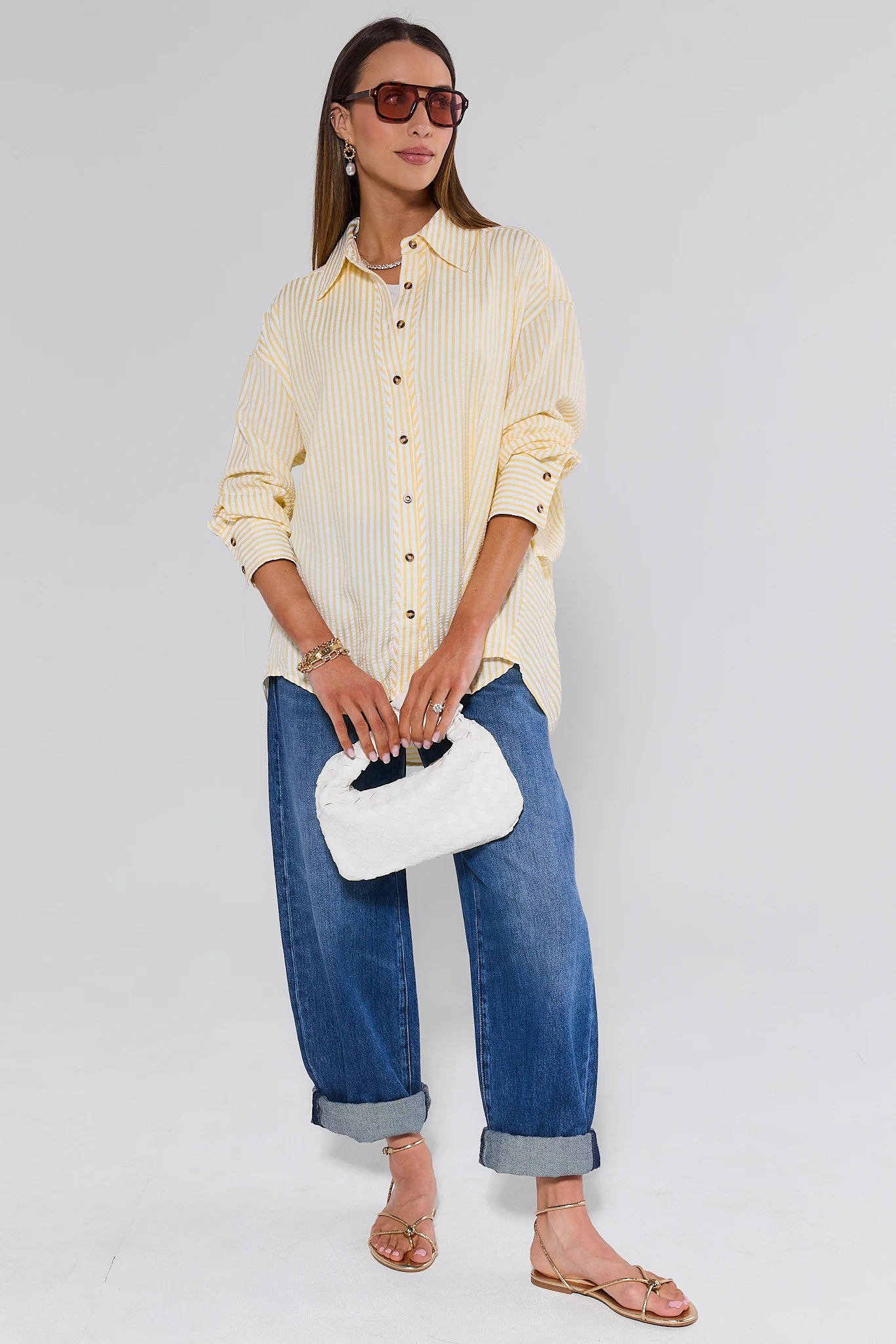 Woman wearing a yellow striped shirt, blue jeans, and holding a white clutch against a plain background