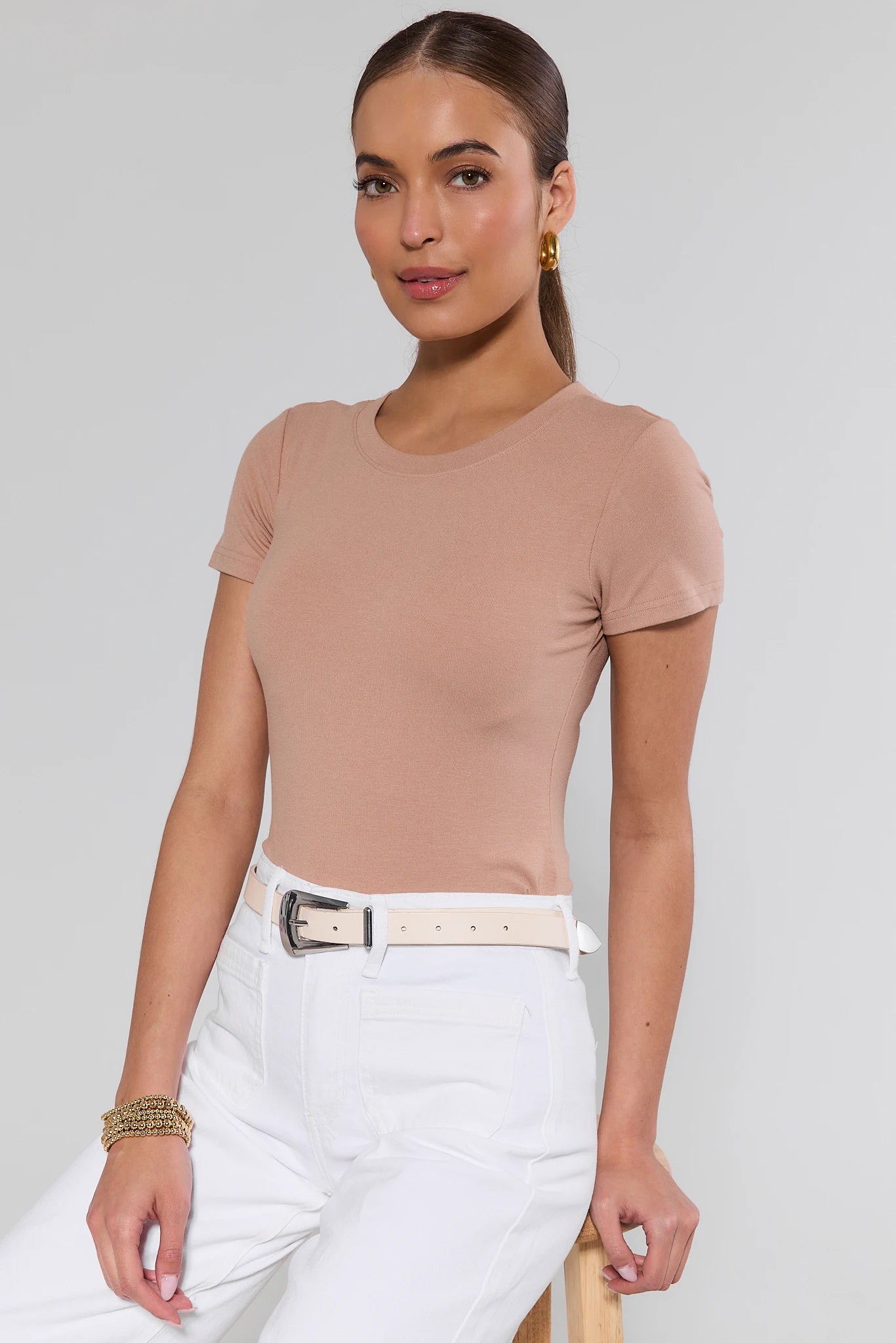 Woman wearing a beige top and white pants with a belt against a plain background