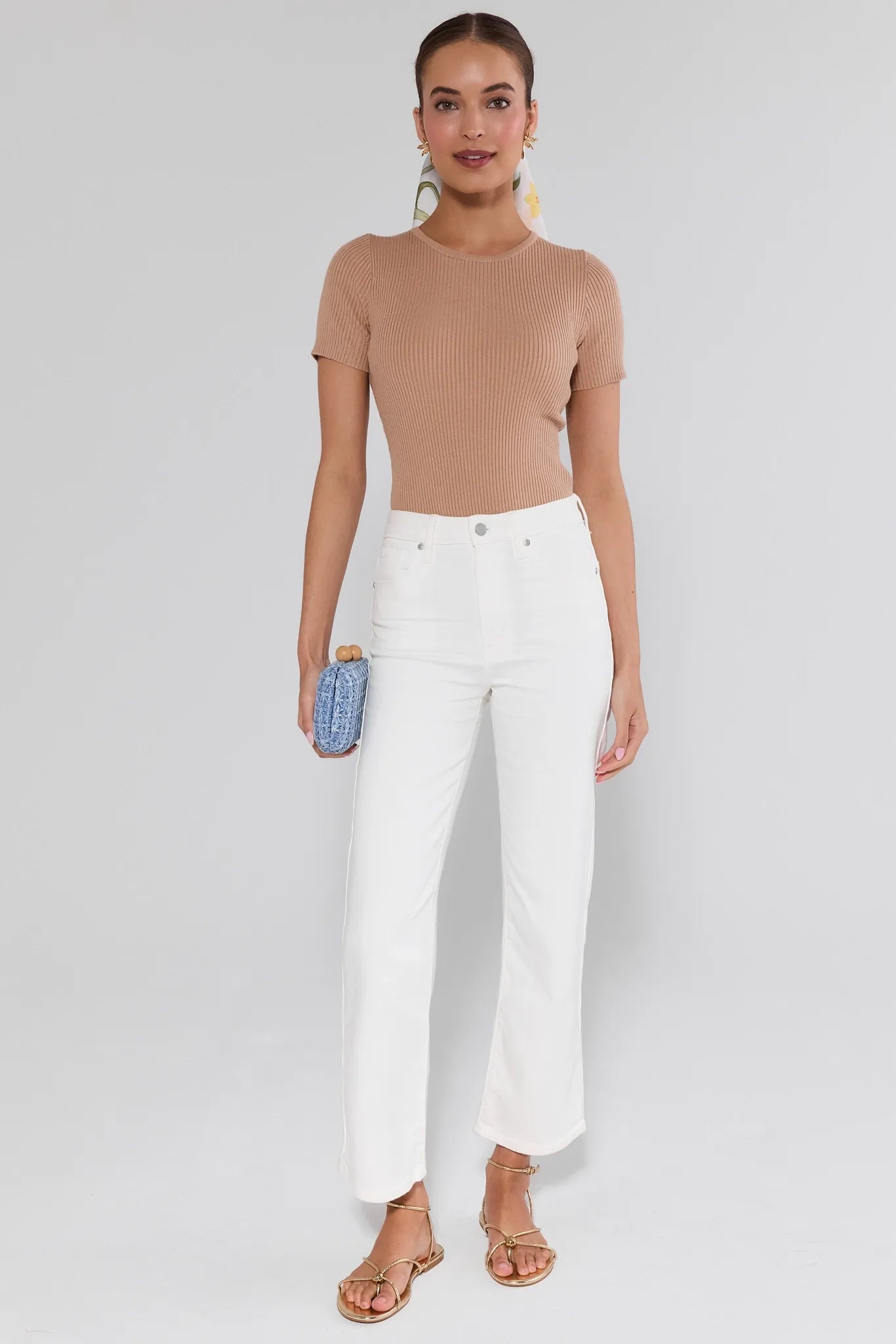 Woman wearing a beige top and white pants with a blue clutch against a white background