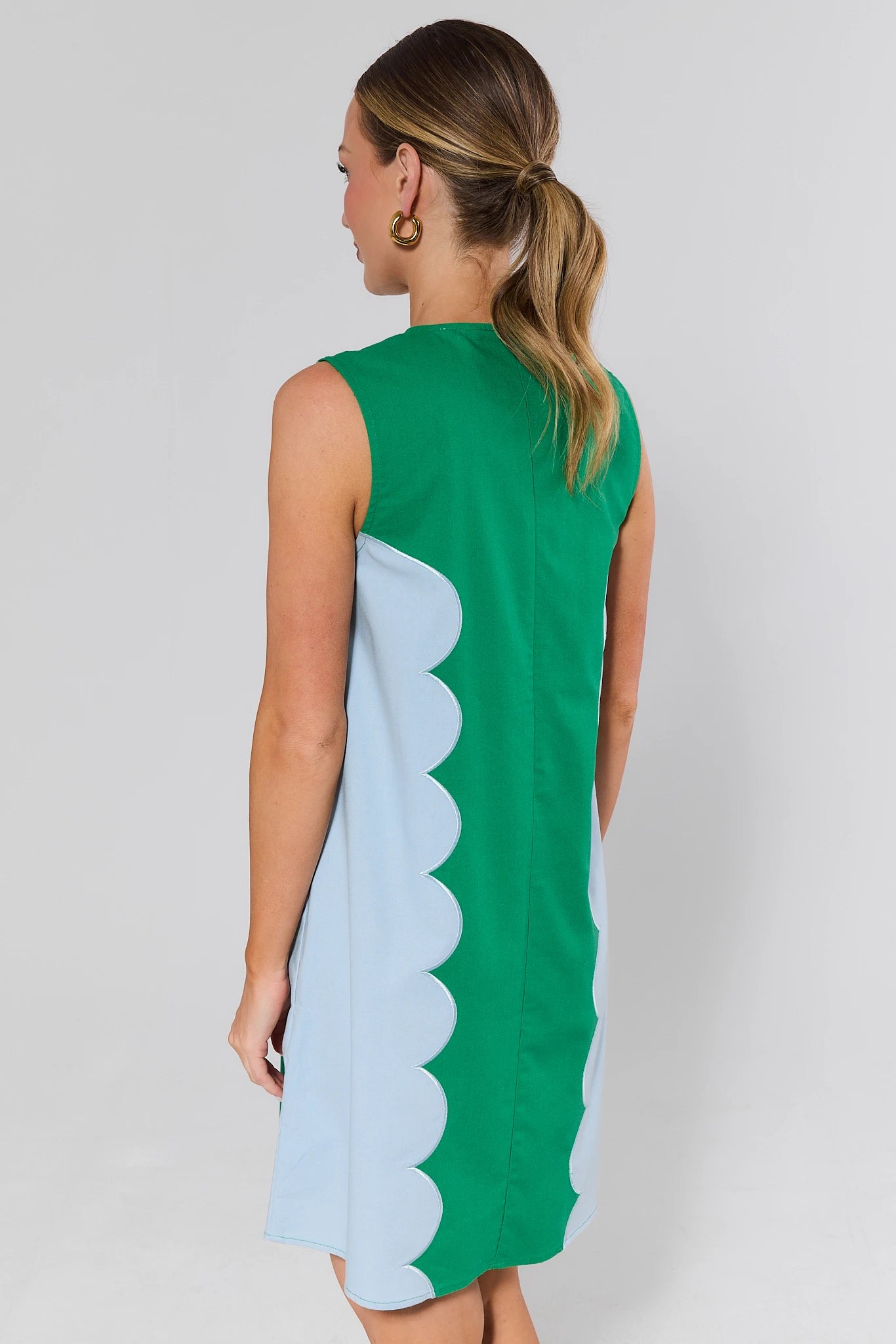 Woman standing wearing a green and light blue dress with a scalloped edge on a plain background
